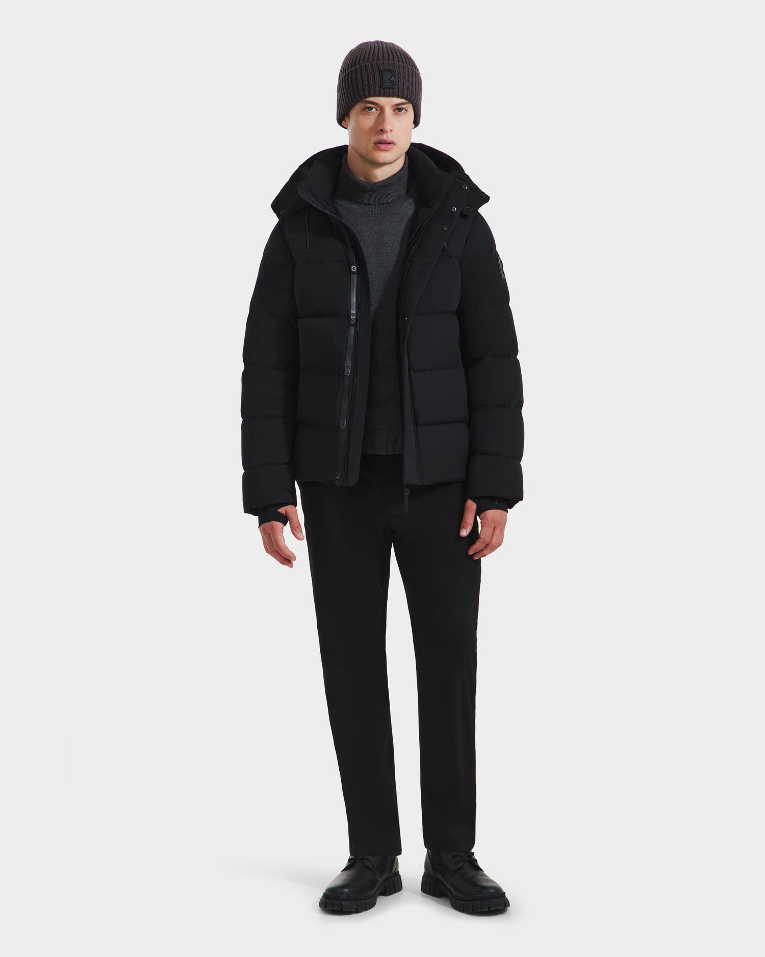 A person faces forward, wearing the Rudsak JOEL - 6124605 Black men’s puffer jacket over a gray sweater, black pants, and black shoes, paired with a gray beanie—showcasing stylish urban outerwear against a plain white background.