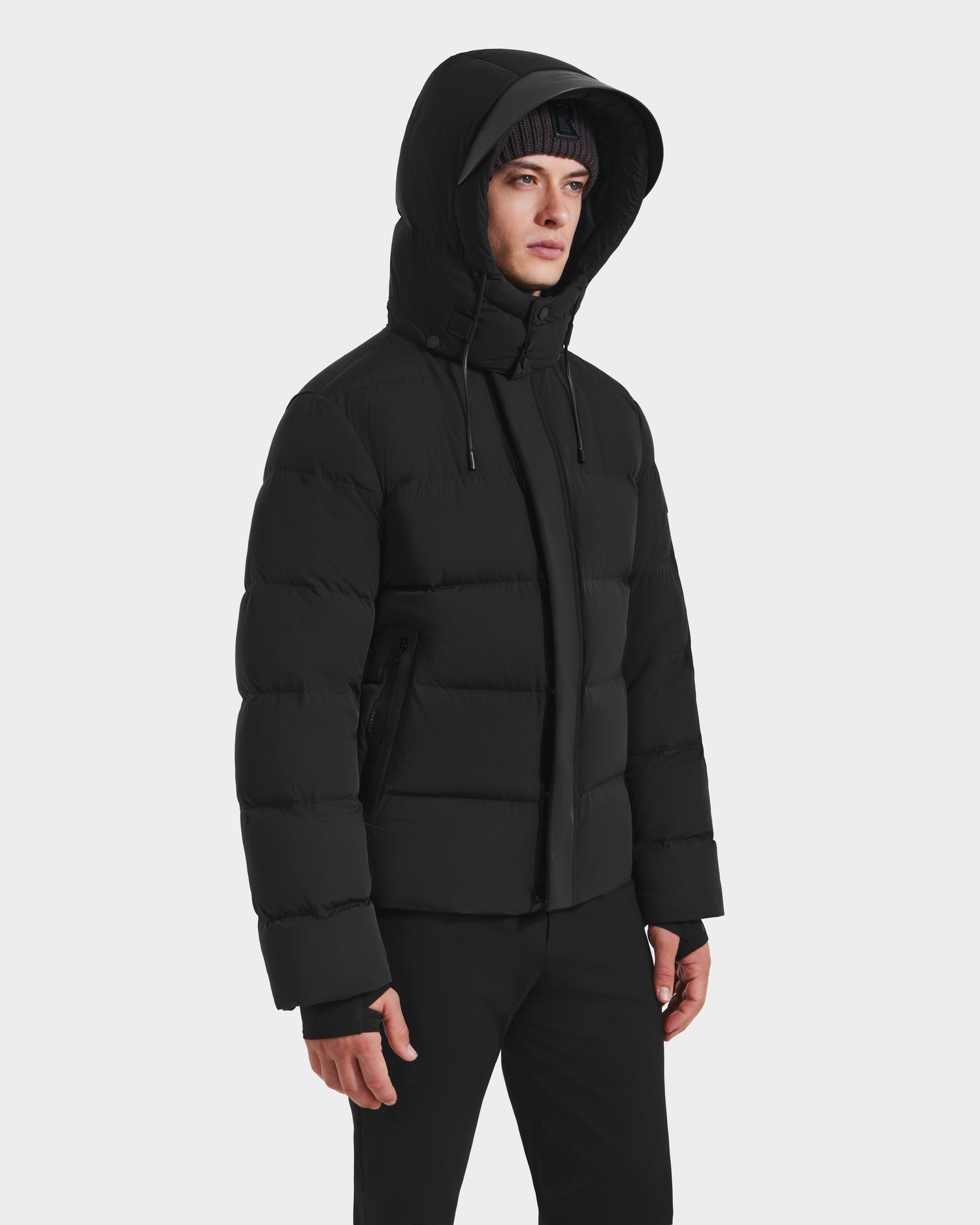 A person in the Rudsak JOEL - 6124605 Black men’s puffer with hood up and black pants stands against a white background, facing slightly to the side.