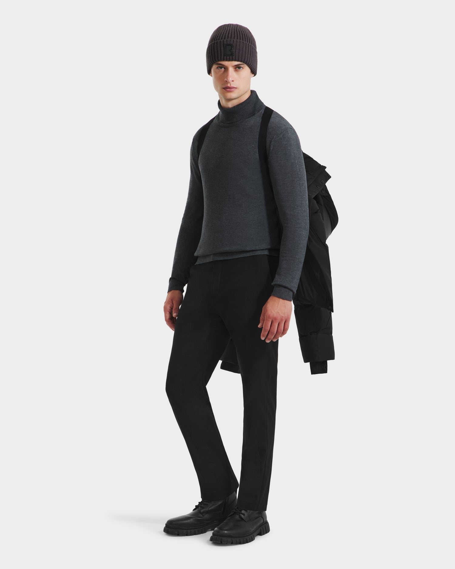 A person stands against a white background with the Rudsak JOEL - 6124605 Black jacket slung over one shoulder, wearing a gray knit beanie, dark gray turtleneck sweater, black pants, and black shoes.