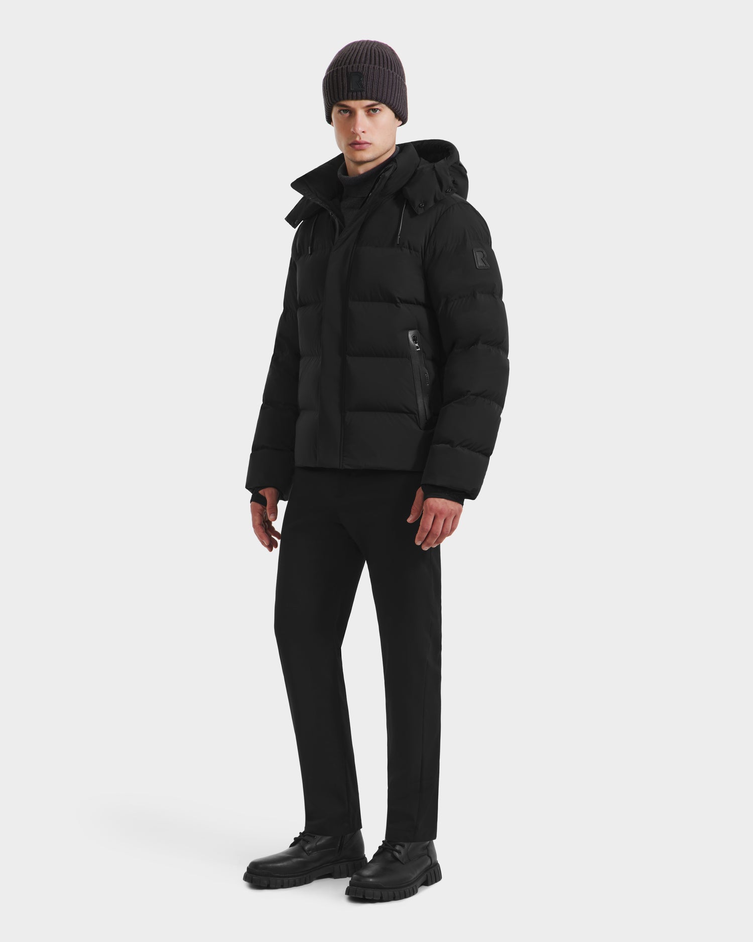 A man stands slightly turned against a plain white background, wearing the Rudsak JOEL - 6124605 Black puffer jacket with matching black pants, shoes, and a dark knit beanie for an urban outerwear-inspired look.