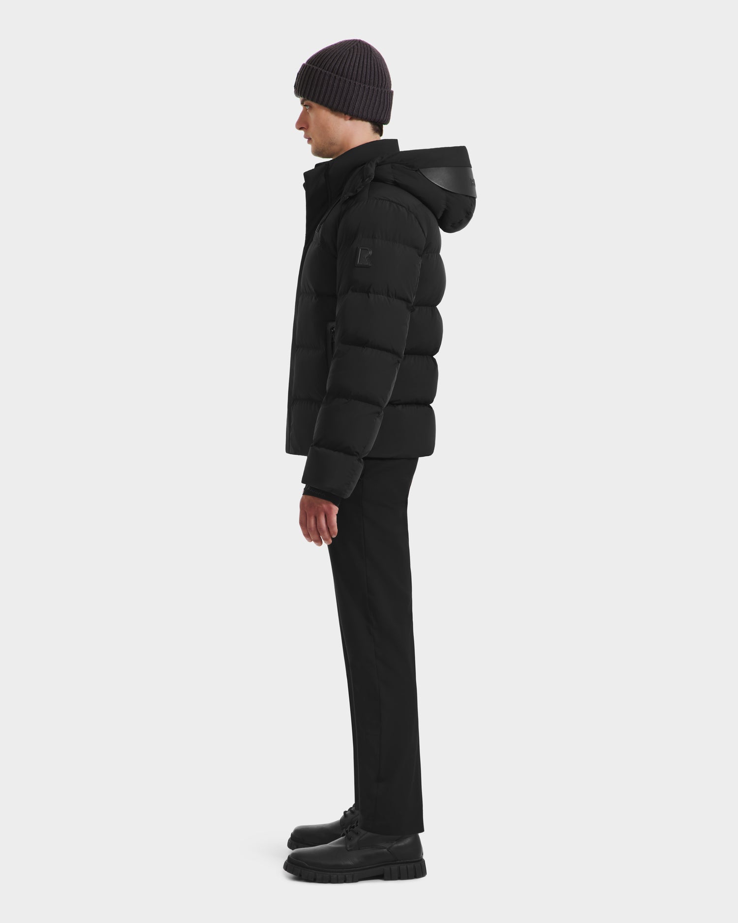 A person stands in profile wearing the Rudsak JOEL - 6124605 Black men’s puffer jacket with hood, paired with black beanie, pants, and shoes—sleek urban outerwear for cold weather—set against a plain white background.