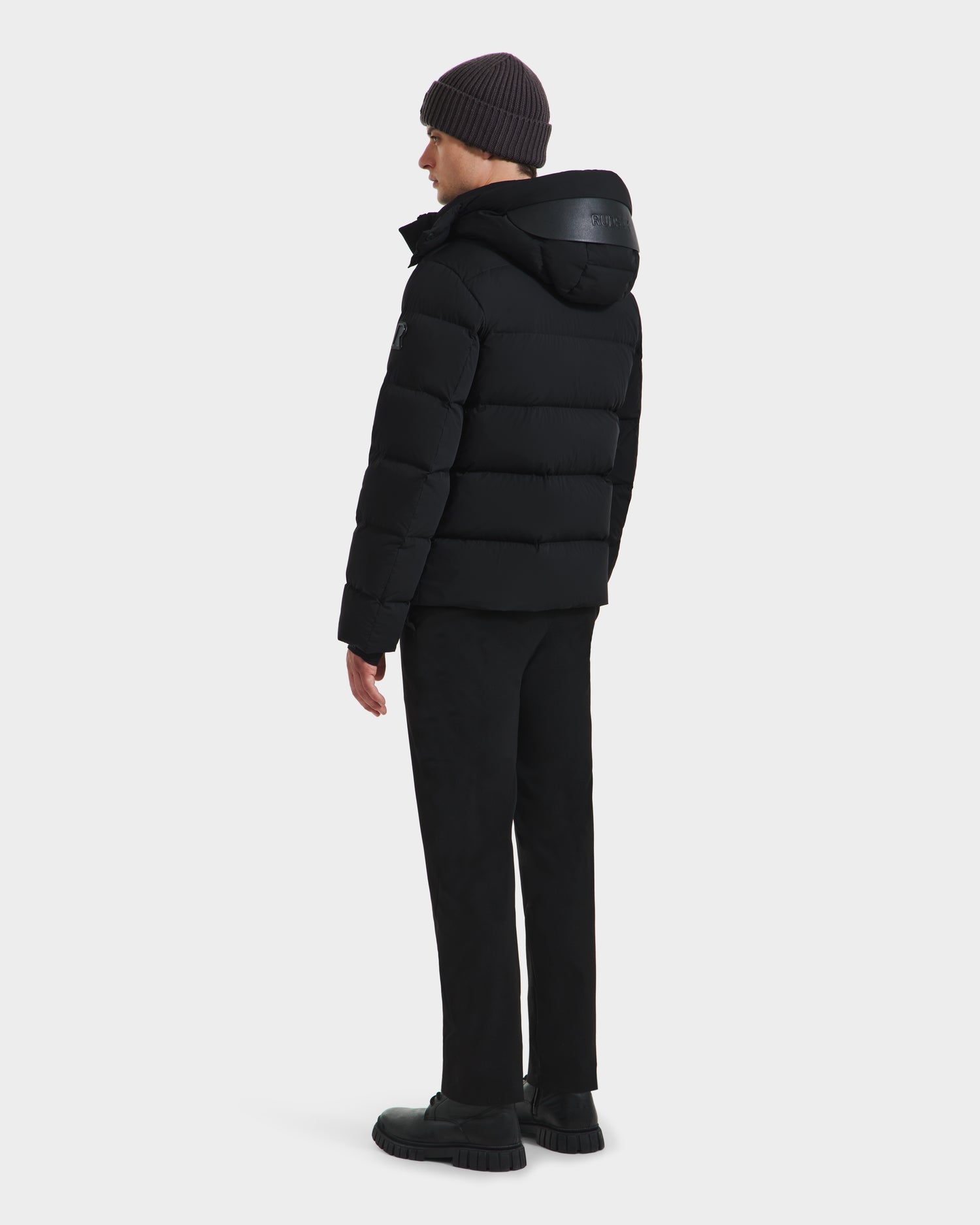 Wearing the Rudsak JOEL - 6124605 Black puffer jacket, dark pants, black shoes, and a dark knit beanie, a man stands with his back turned and glances to the side against a plain light background.