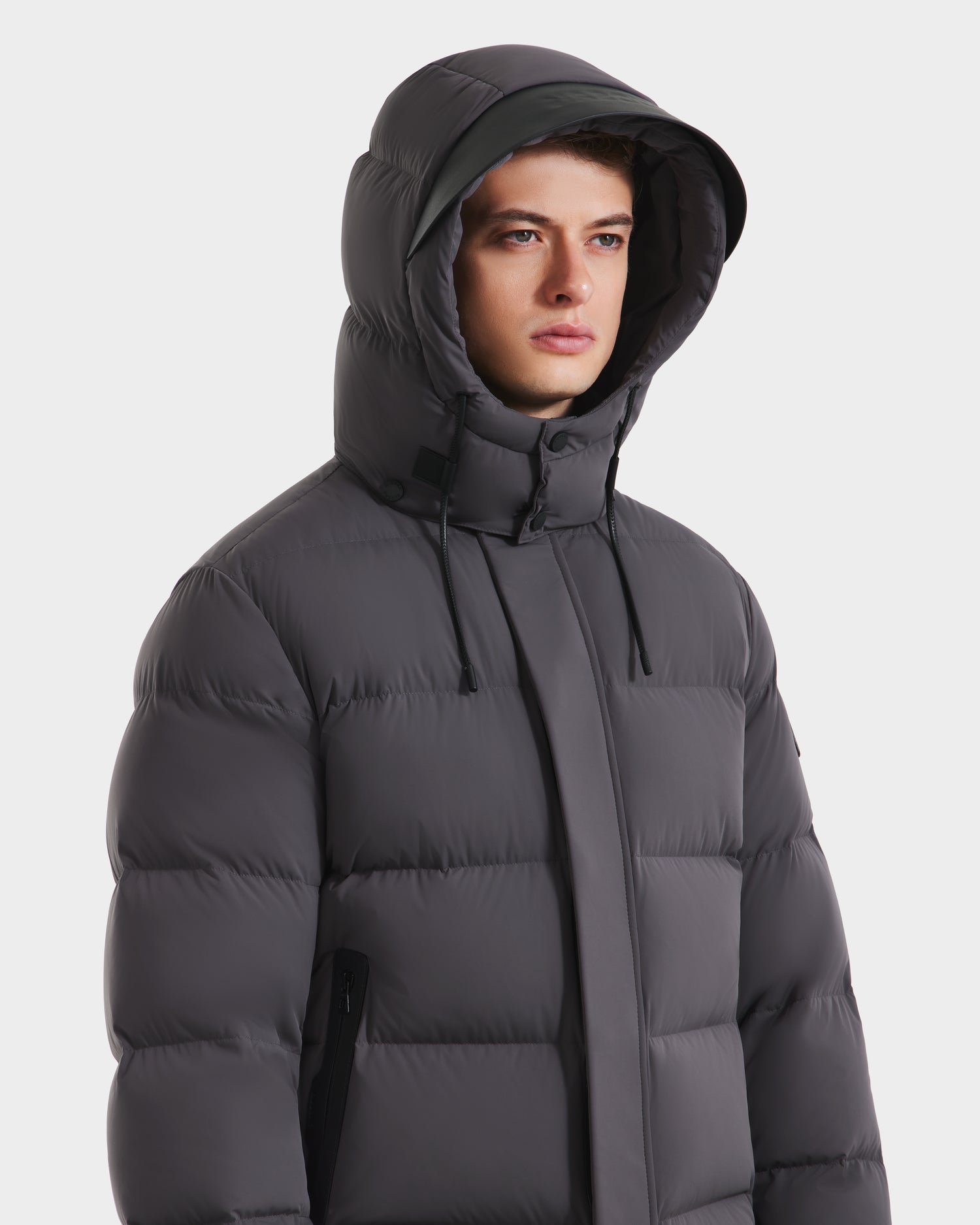 A person wearing the Rudsak JOEL - 6124605 Grey men’s puffer with a weather-resistant shell and hood, looking slightly to the side against a plain white background.