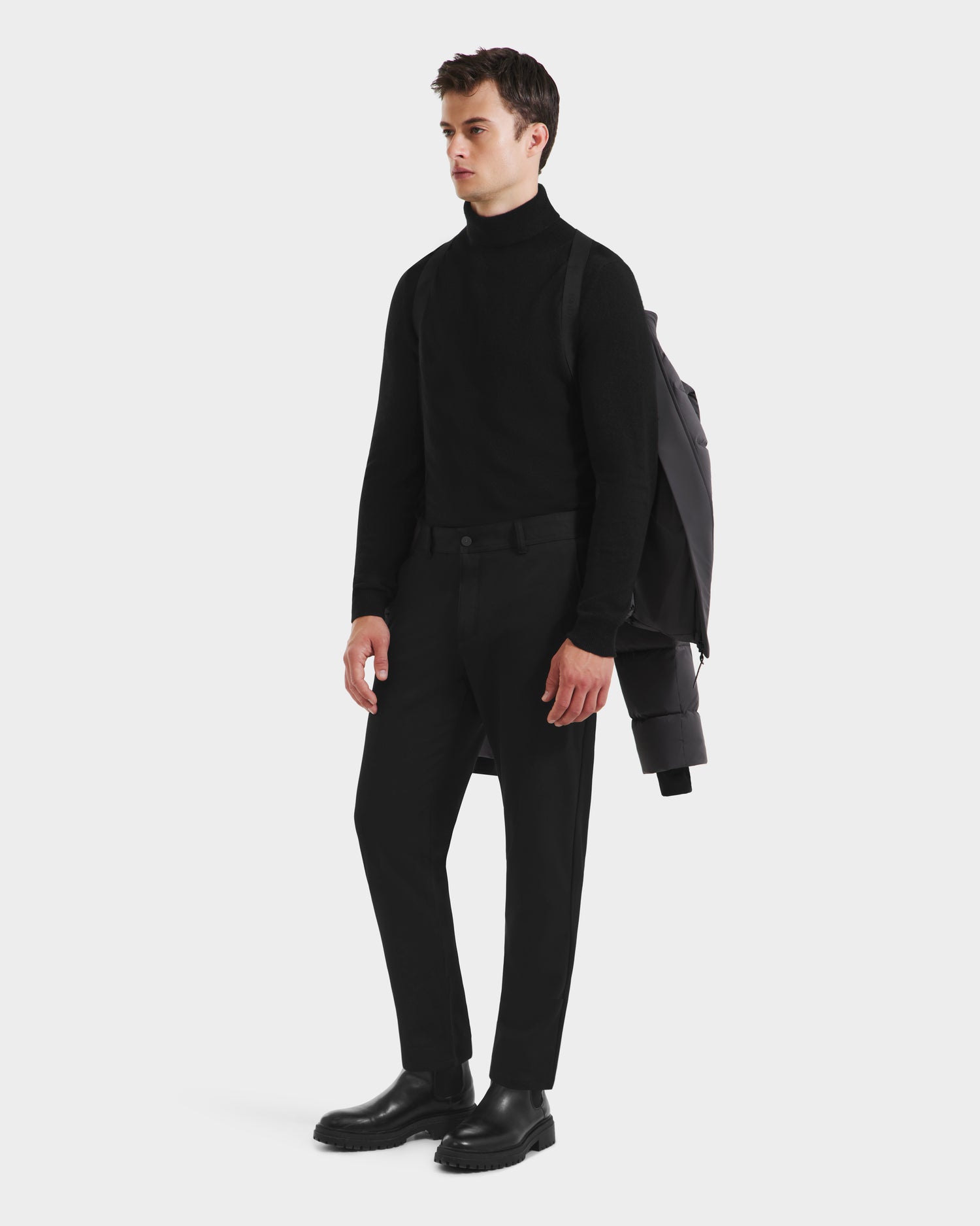 A man stands against a plain background wearing a black turtleneck, black pants, and black boots, with the Rudsak JOEL - 6124605 Grey weather-resistant shell jacket draped over his shoulders.