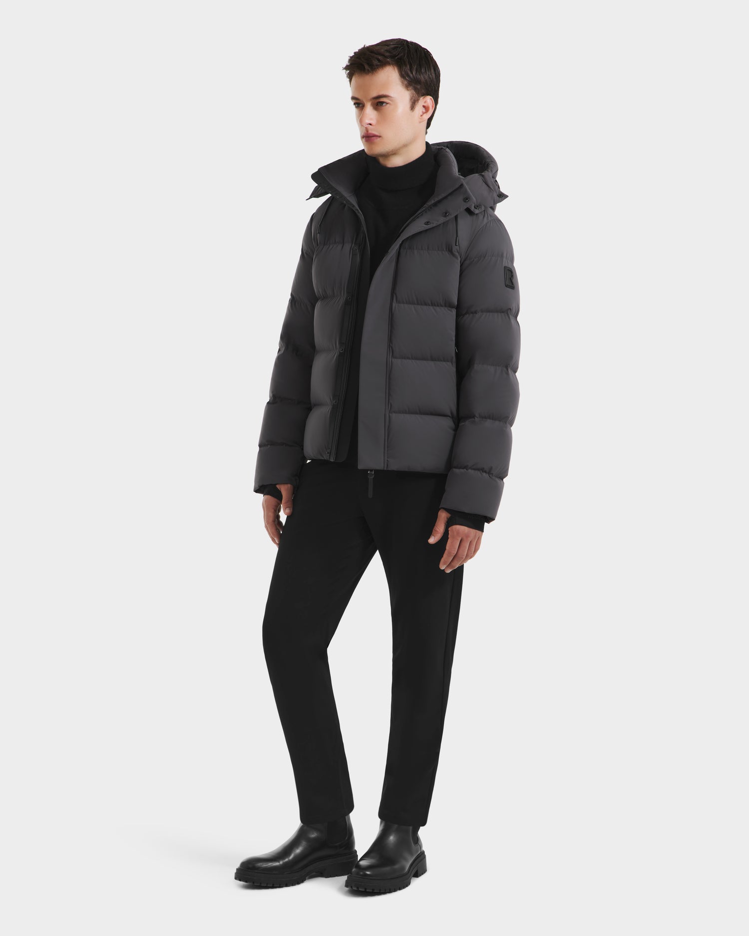 A young man stands against a plain white background wearing the Rudsak JOEL - 6124605 Grey men’s puffer with weather-resistant shell, paired with a black turtleneck, black pants, and black boots.