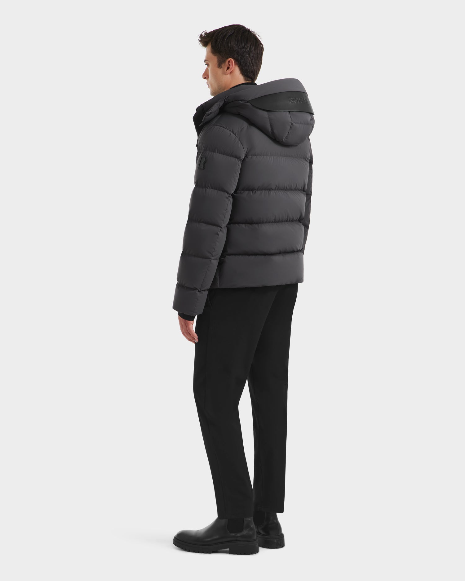 A man faces away, wearing the Rudsak JOEL - 6124605 Grey men's puffer with high-loft down fill and weather-resistant shell, paired with black pants and boots, set against a plain white background.