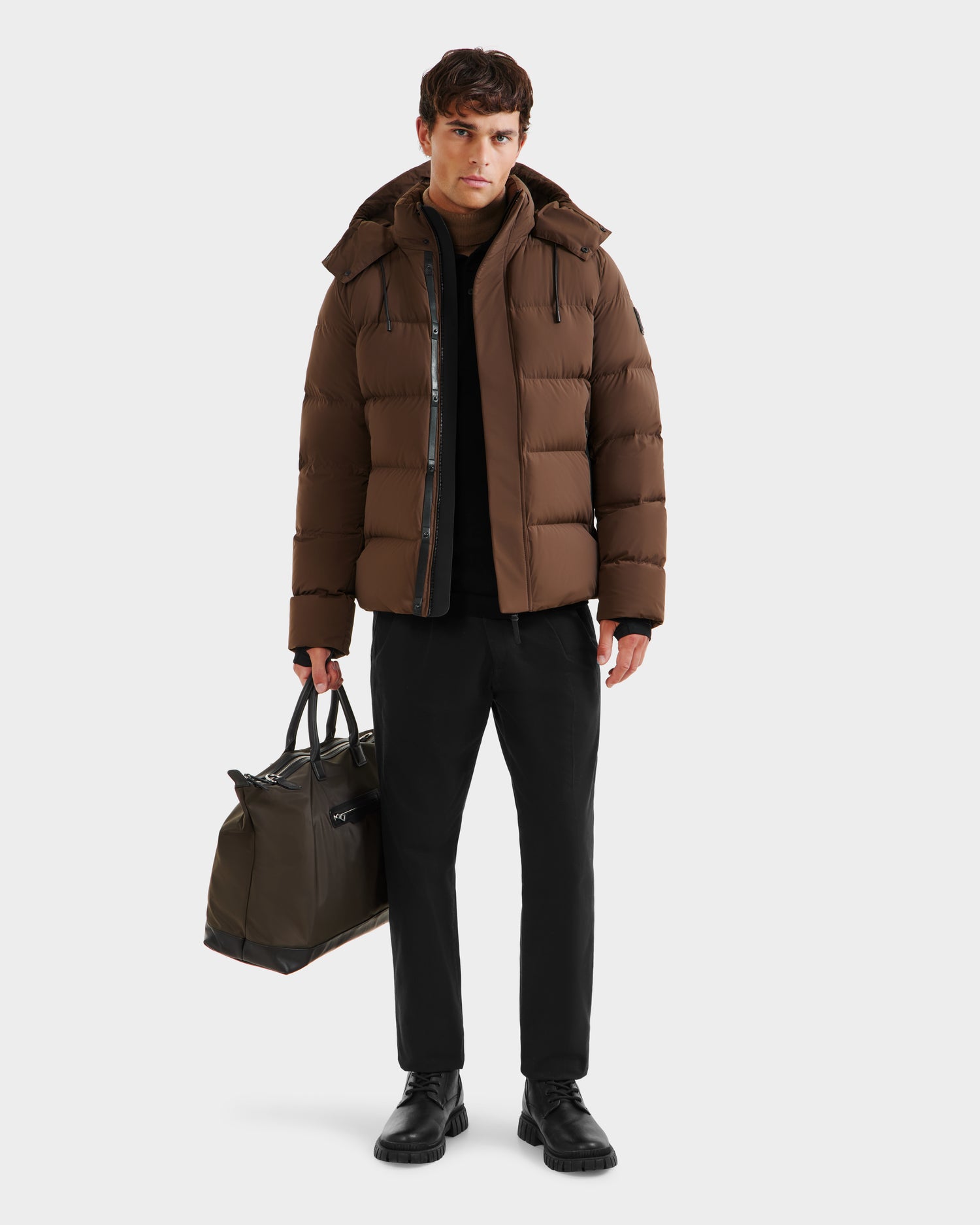 A man stands against a plain white background, holding a dark green duffel bag and wearing Rudsak’s JOEL - 6124605 Mocha men’s puffer jacket, black pants, and black shoes, showcasing cold-weather performance and weather-resistant style.