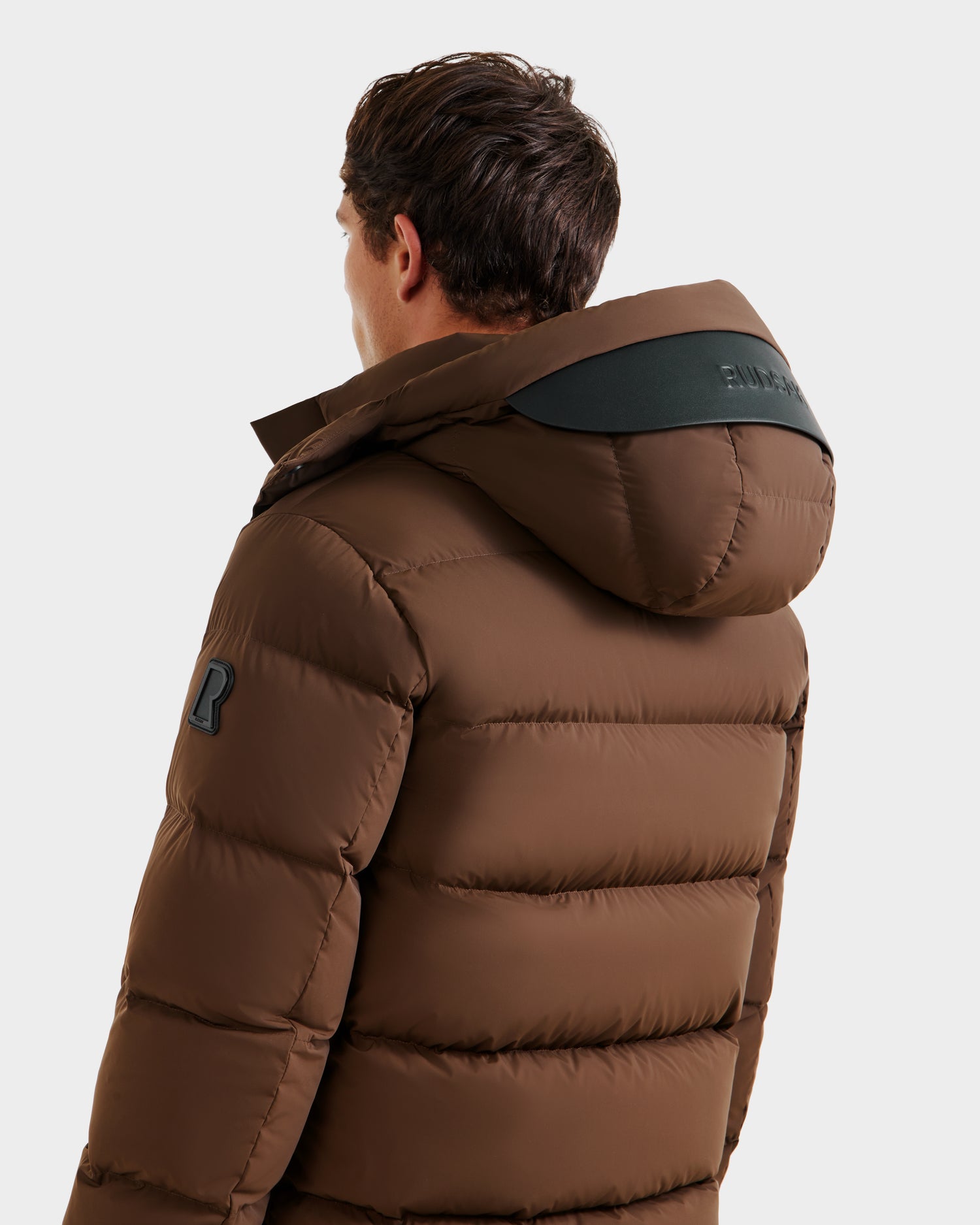 A person is seen from the back wearing the Rudsak JOEL - 6124605 Mocha men’s puffer jacket with a hood and black patch details on the shoulder and sleeve, ideal for cold-weather performance.