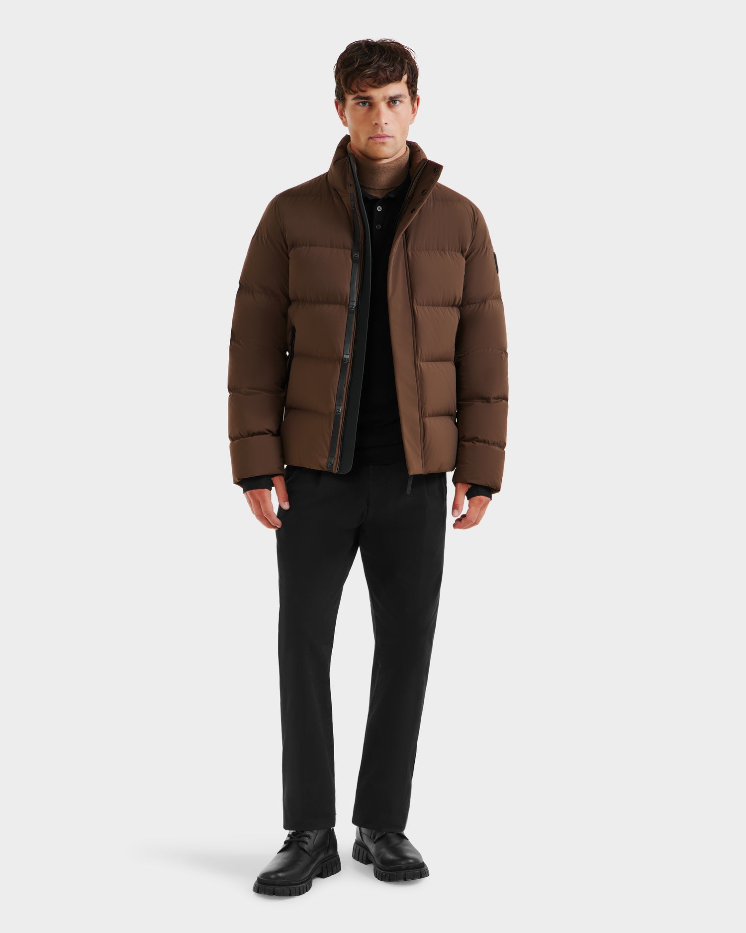 A man stands against a plain background wearing the Rudsak JOEL - 6124605 Mocha men’s puffer jacket, black pants, and black shoes. He faces forward with a neutral expression, hands relaxed at his sides, ready for cold-weather performance.