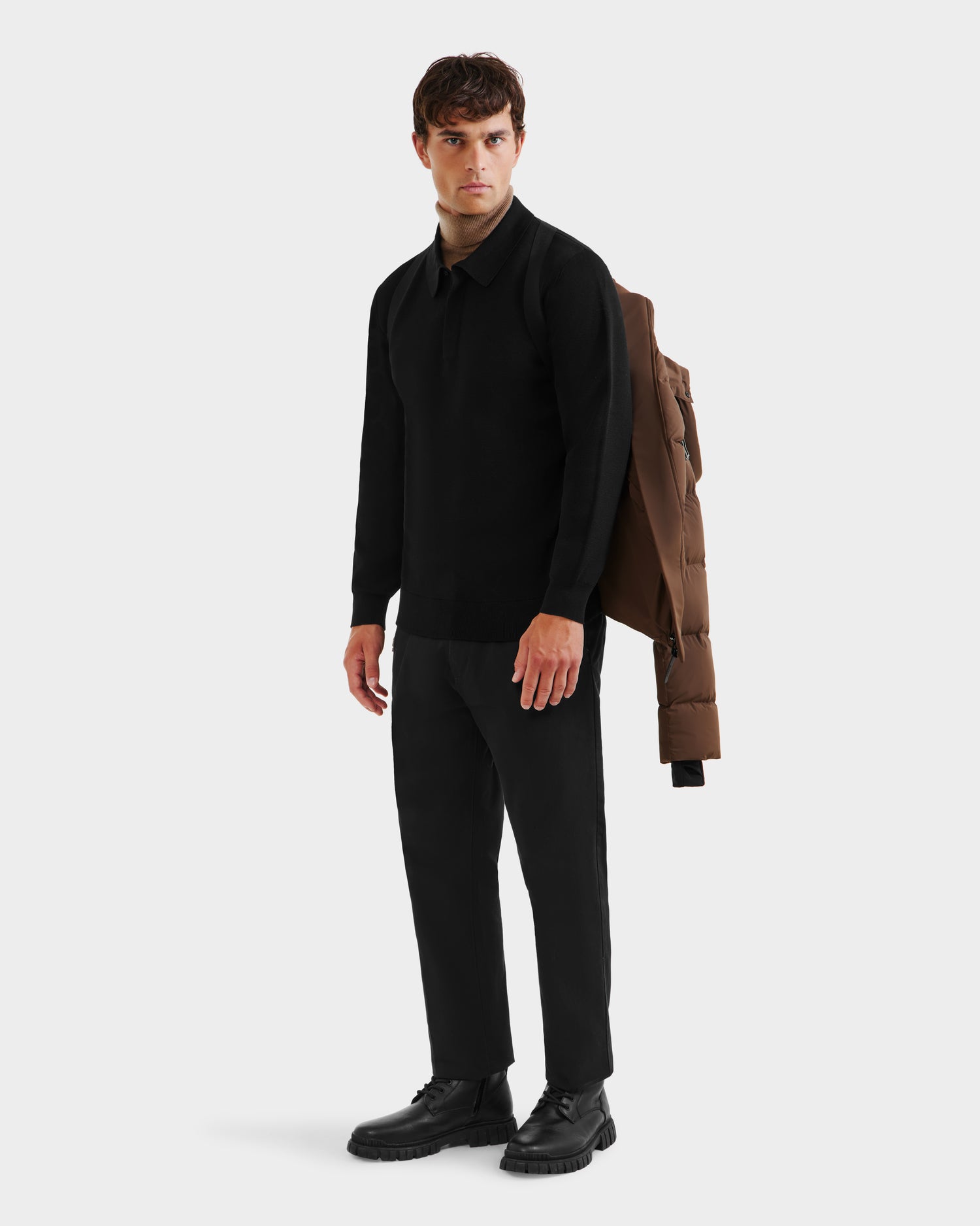 A man stands against a plain background in a black long-sleeve shirt and pants, with the Rudsak JOEL - 6124605 Mocha men’s puffer jacket draped over his shoulder—highlighting its versatile cold-weather performance and modern style.
