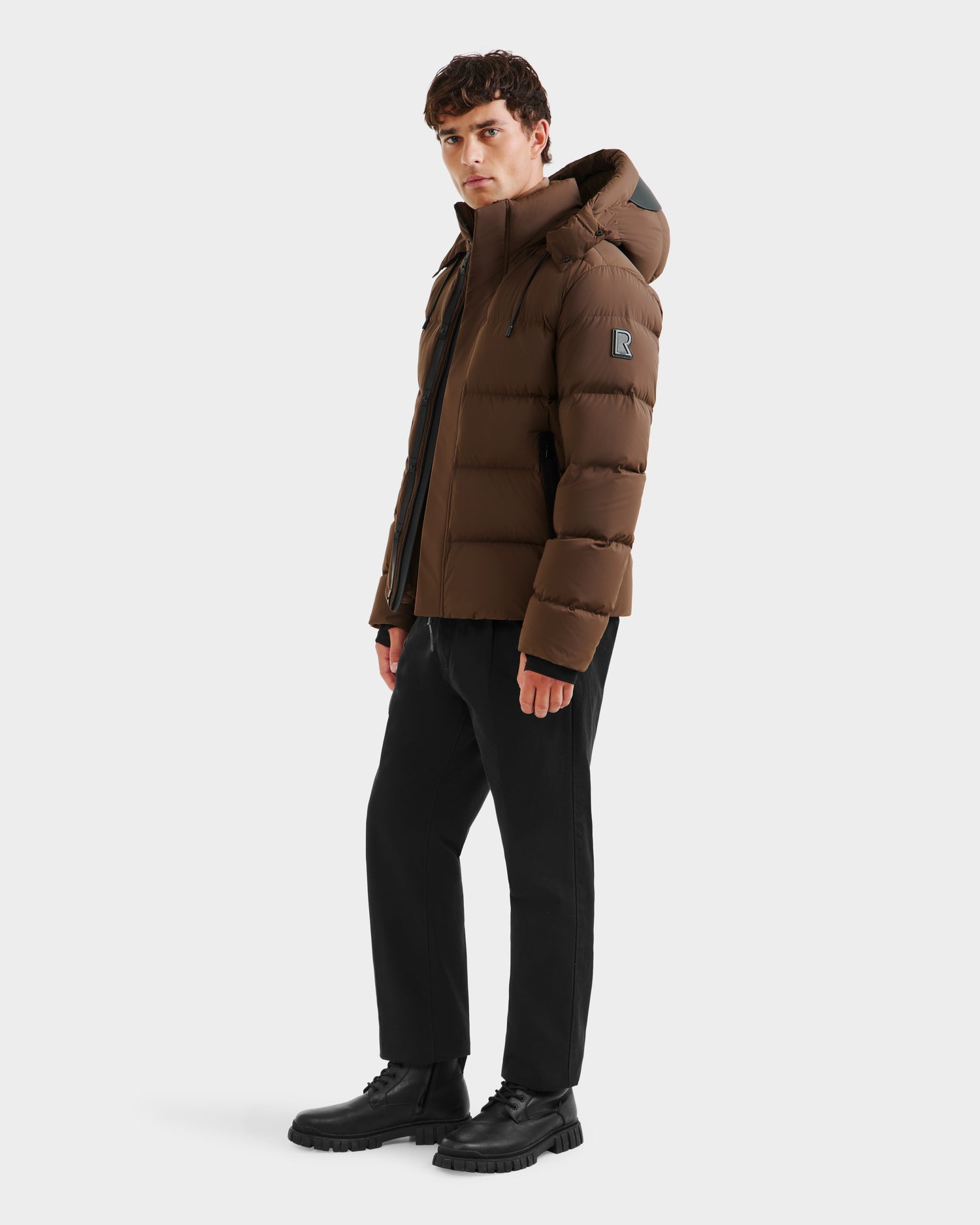 A man wears the Rudsak JOEL - 6124605 Mocha weather-resistant mens puffer jacket with a hood, paired with black pants and boots, standing against a plain white background and looking slightly to his left.