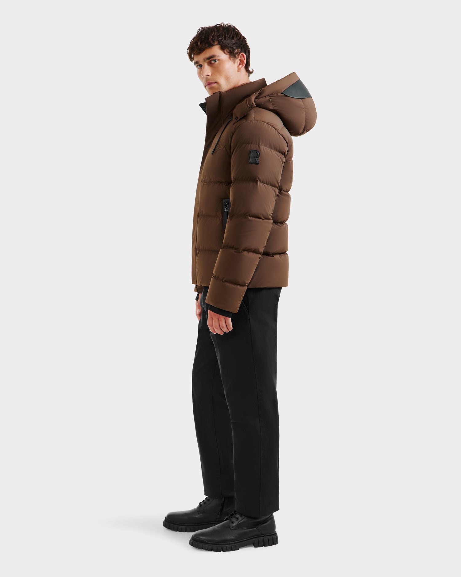 A man stands sideways against a plain white background wearing the Rudsak JOEL - 6124605 Mocha weather-resistant mens puffer jacket with a hood, paired with black pants and black shoes.