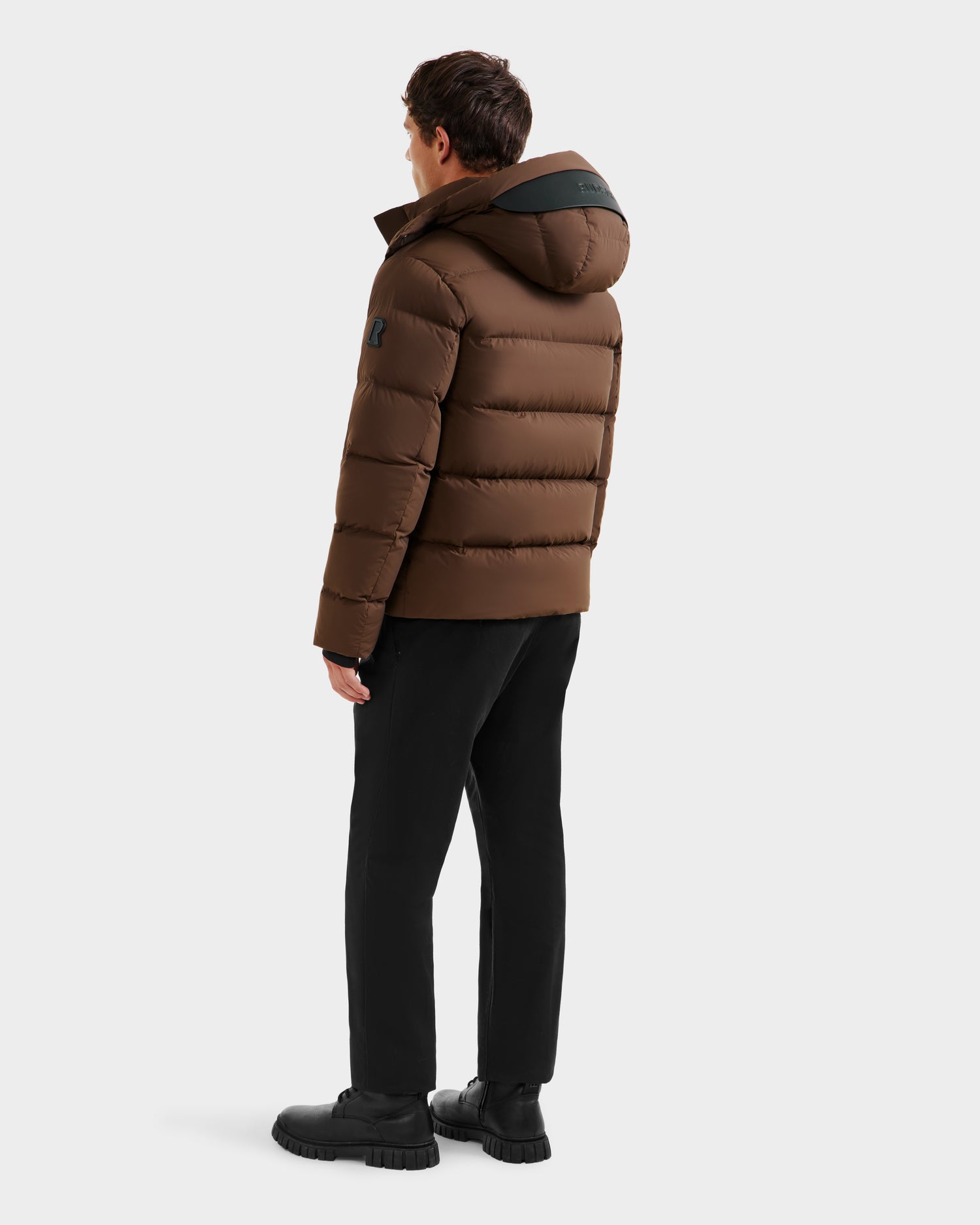 A man faces away, wearing the Rudsak JOEL - 6124605 Mocha men’s puffer down jacket with a hood, paired with black pants and shoes. The plain, light-toned background highlights this cold-weather performance style.