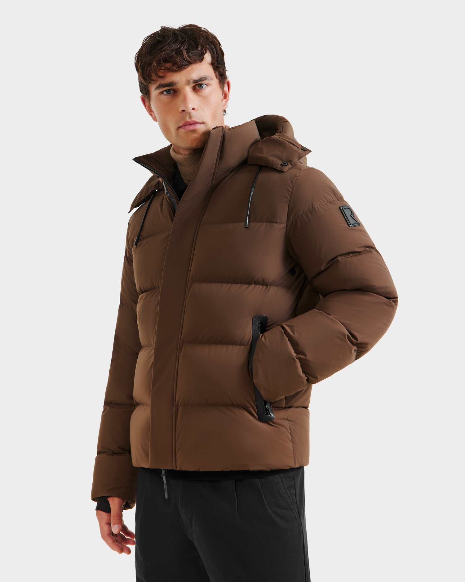 A man wearing the Rudsak JOEL - 6124605 Mocha weather-resistant men’s puffer jacket with a hood and black zipper pockets stands against a plain light background, looking to the side with one hand in his pocket.