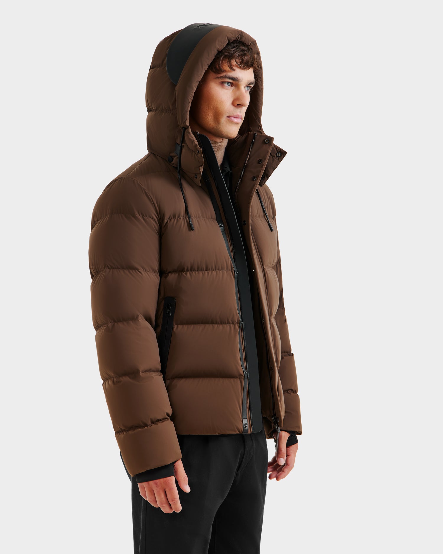 A person models the Rudsak JOEL - 6124605 Mocha men’s puffer jacket in brown with black zipper accents and a hood, designed for cold-weather performance. It’s styled with black pants against a plain, light background.