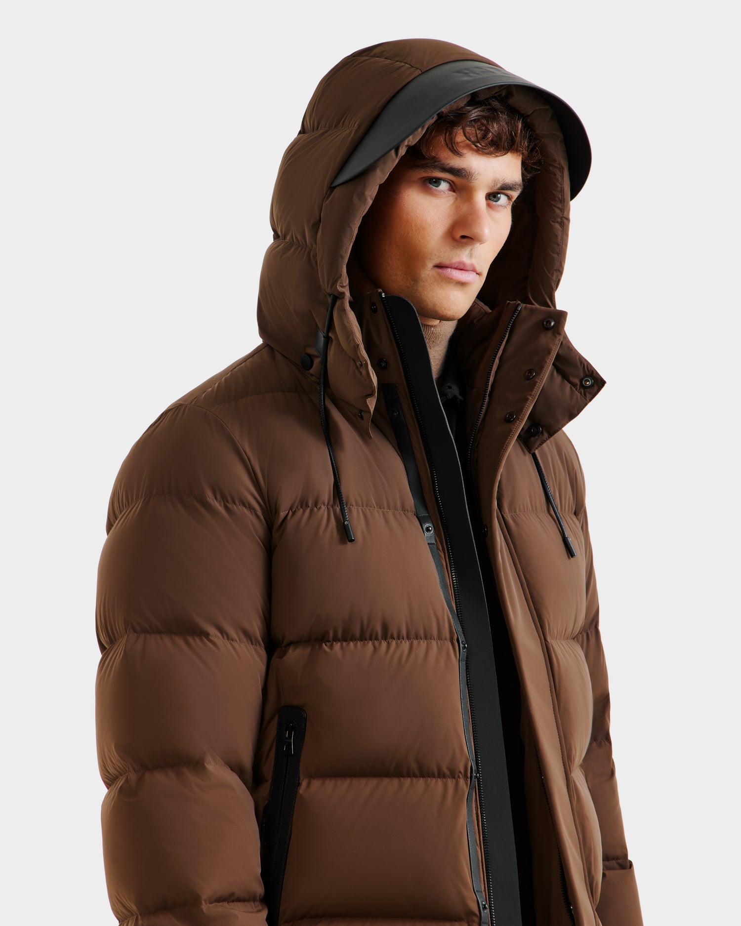 A person in the Rudsak JOEL - 6124605 Mocha mens puffer jacket with hood and black zipper looks slightly to the side, highlighting its cold-weather performance against a plain, light background.