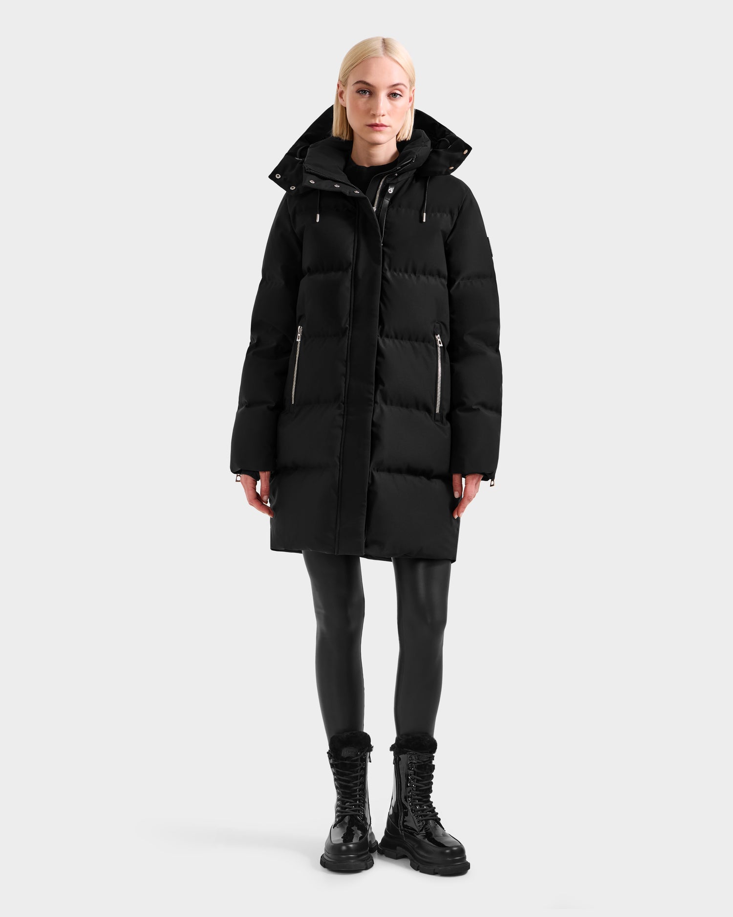 A woman with straight blonde hair wears the Rudsak JOELLE H - 8125602 Black women’s down puffer coat with a hood, paired with black leggings and black lace-up boots, standing against a plain light background.