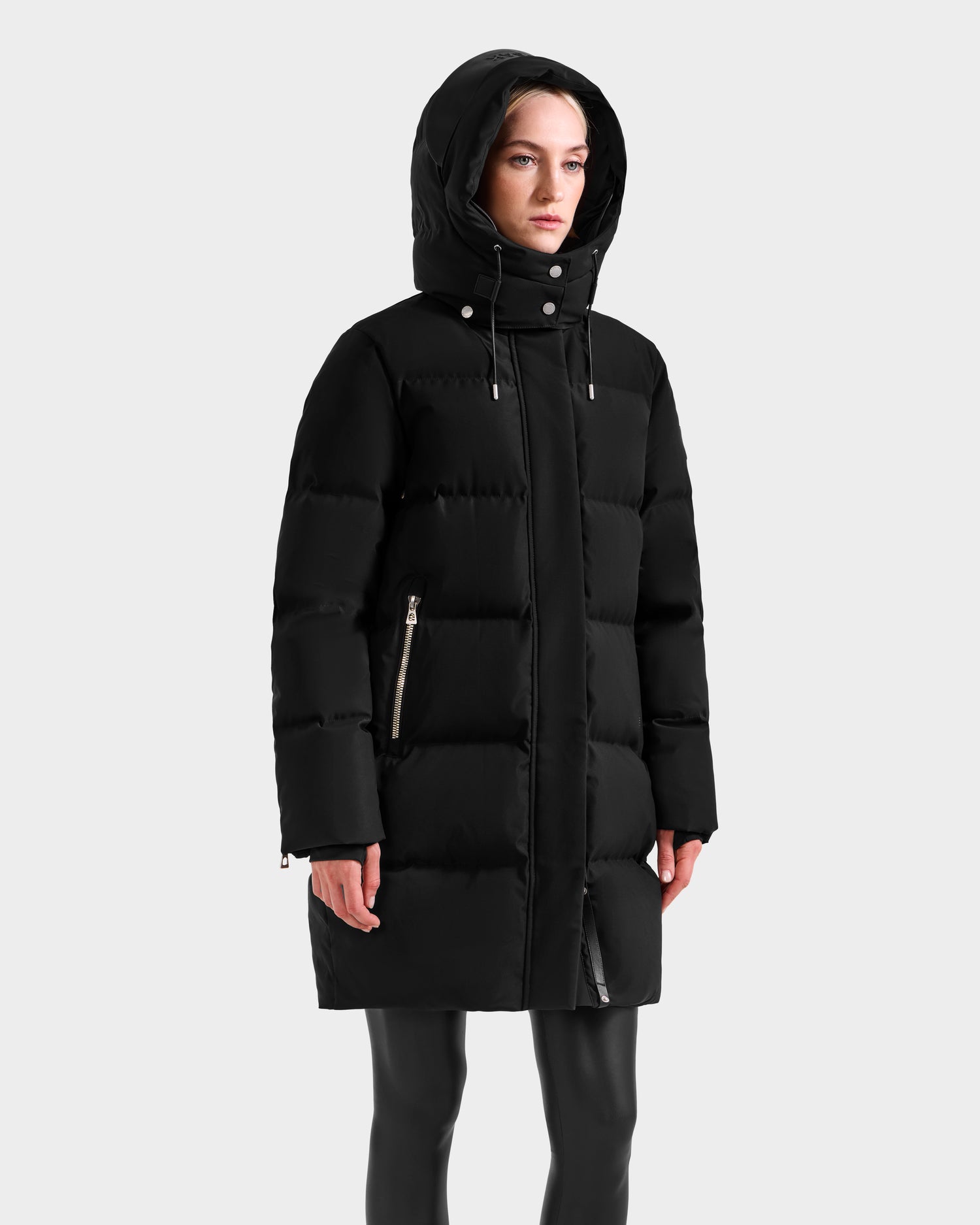 A person models the Rudsak JOELLE H - 8125602 Black, a women's knee-length down puffer coat with hood, front zipper, and side pockets, standing against a plain light gray background.