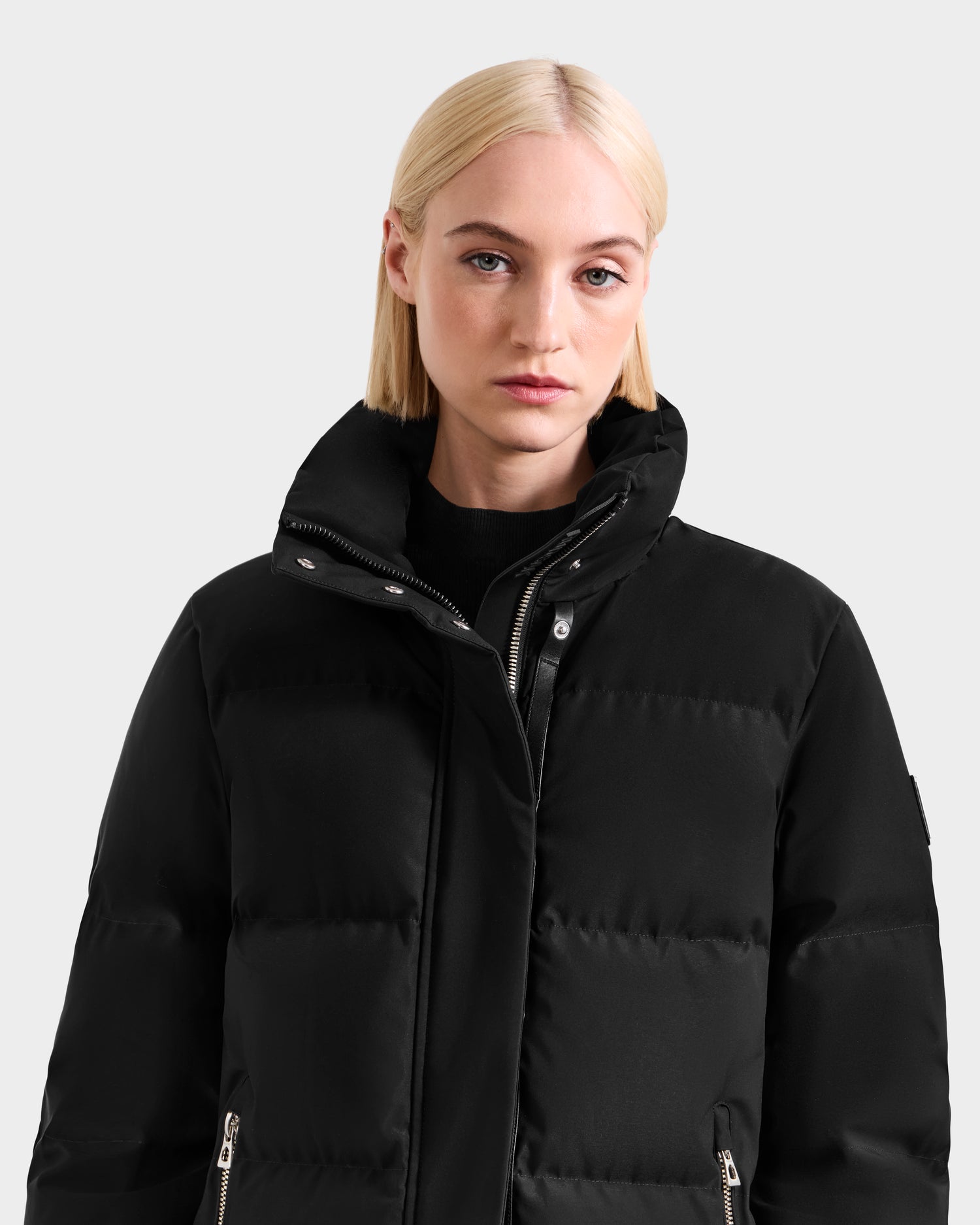 A woman with straight, shoulder-length blonde hair models the Rudsak JOELLE H - 8125602 black women's down puffer jacket with a high collar, standing against a plain light gray background.