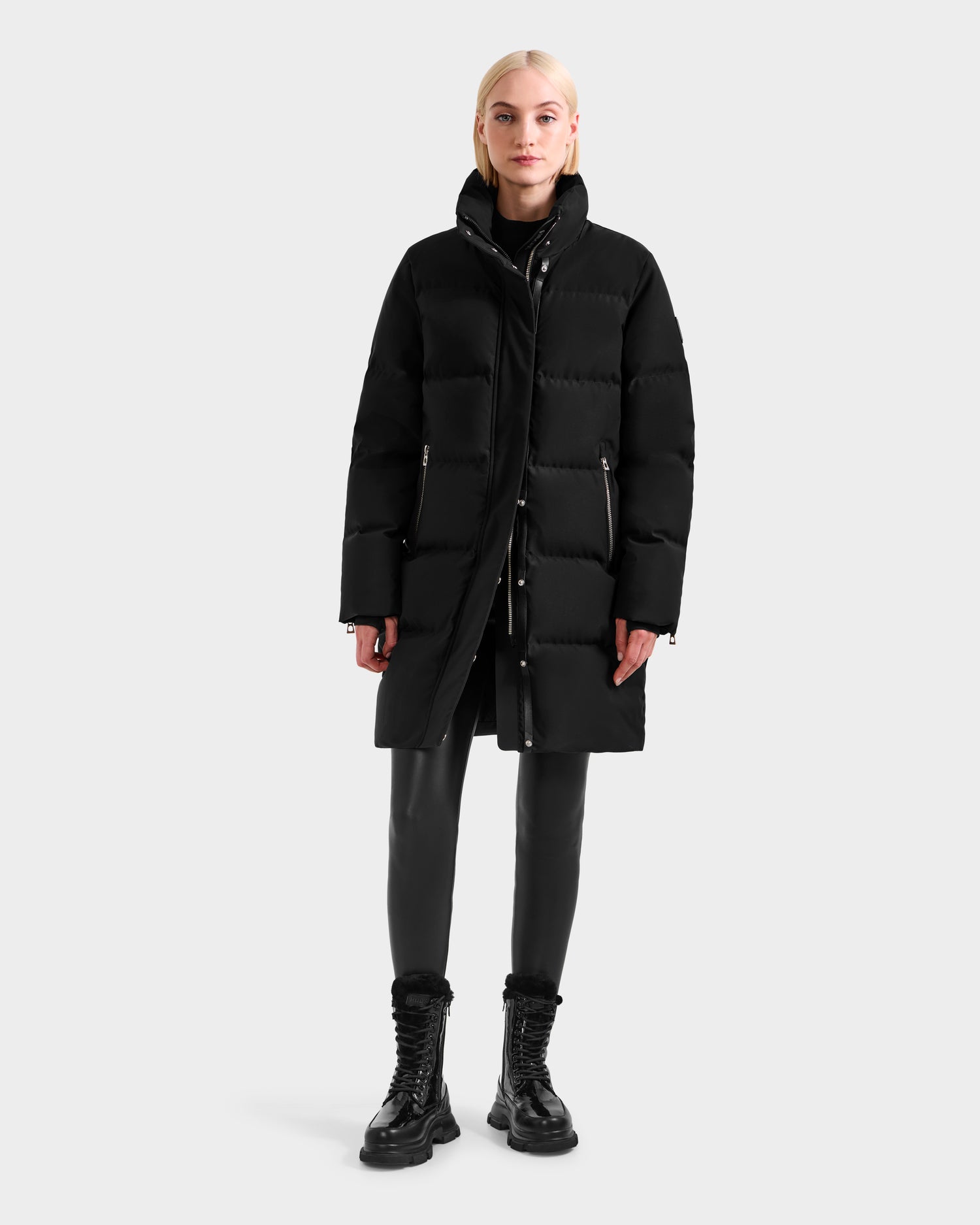 A woman faces forward in the Rudsak JOELLE H - 8125602 Black down puffer coat, styled with black leggings and lace-up boots. Her blond hair is tucked behind her ears as she poses against a plain light gray background.