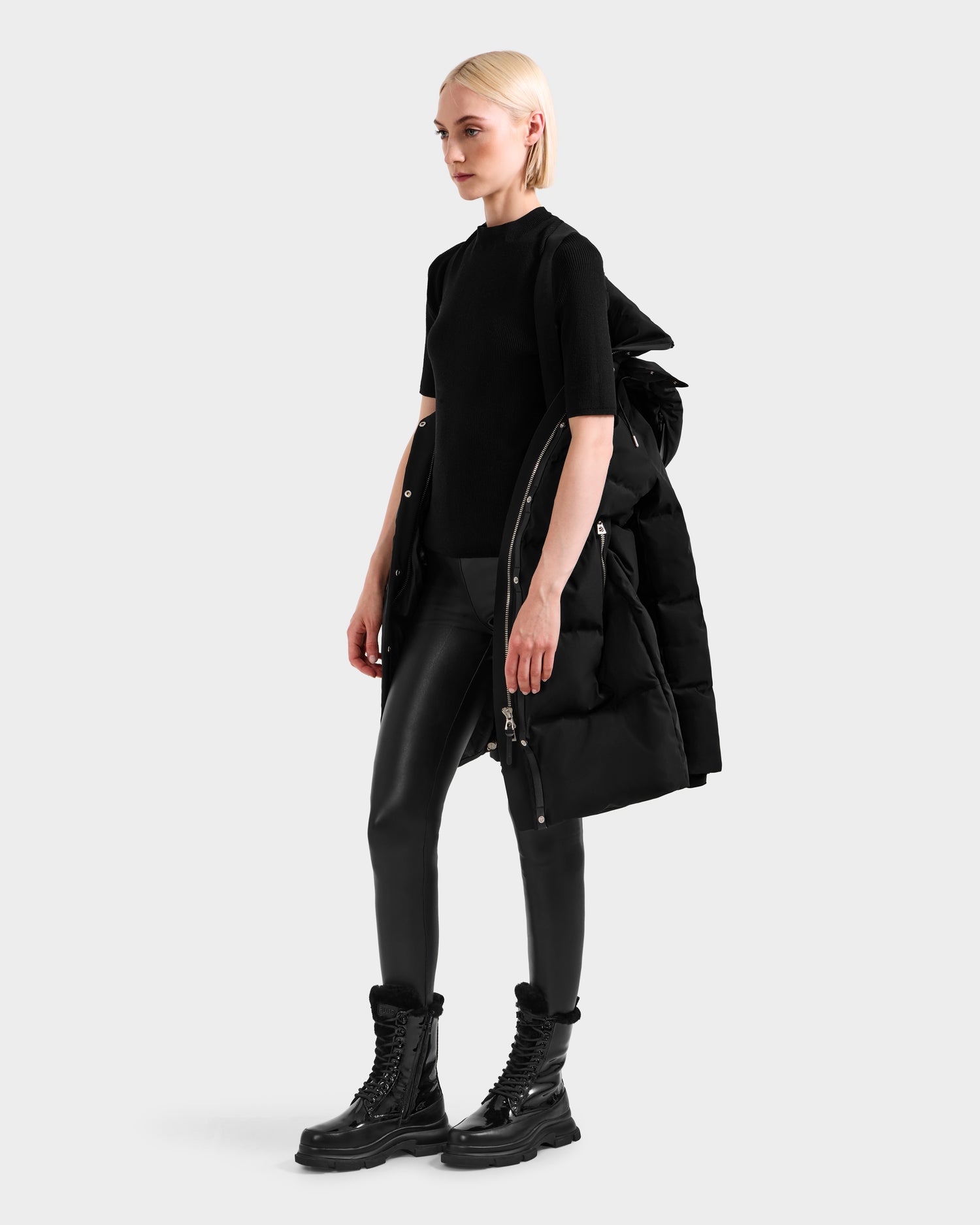 A blonde woman poses against a light gray background, draping the Rudsak JOELLE H - 8125602 Black women’s down puffer over one shoulder, styled with a black top, leather pants, and combat boots for a chic, water-resistant winter look.
