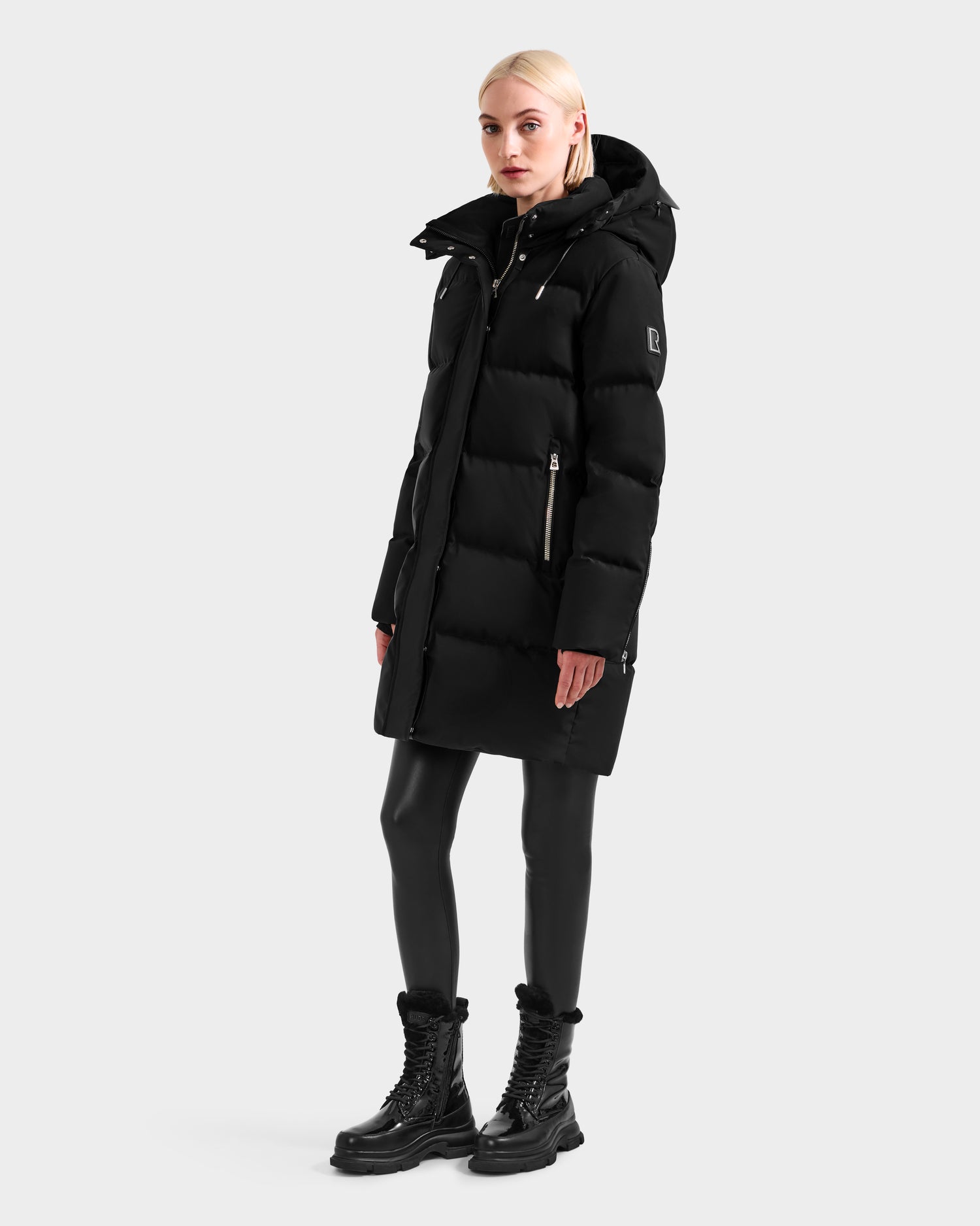 A blonde woman wears the Rudsak JOELLE H - 8125602 Black women's down puffer coat with black leggings and lace-up boots, standing against a plain light gray background.