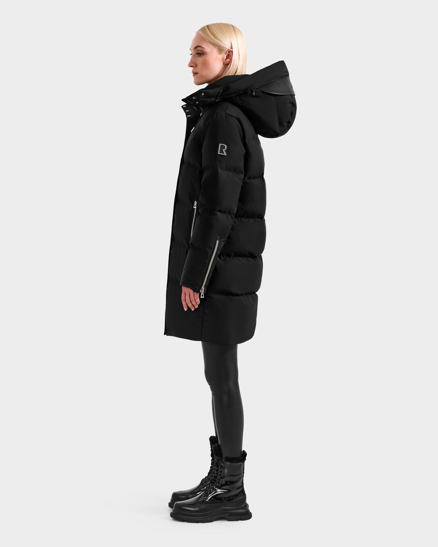 A person with straight blond hair stands sideways, wearing the Rudsak JOELLE H - 8125602 Black women’s down puffer coat with a hood, paired with black leggings and boots against a plain light background.