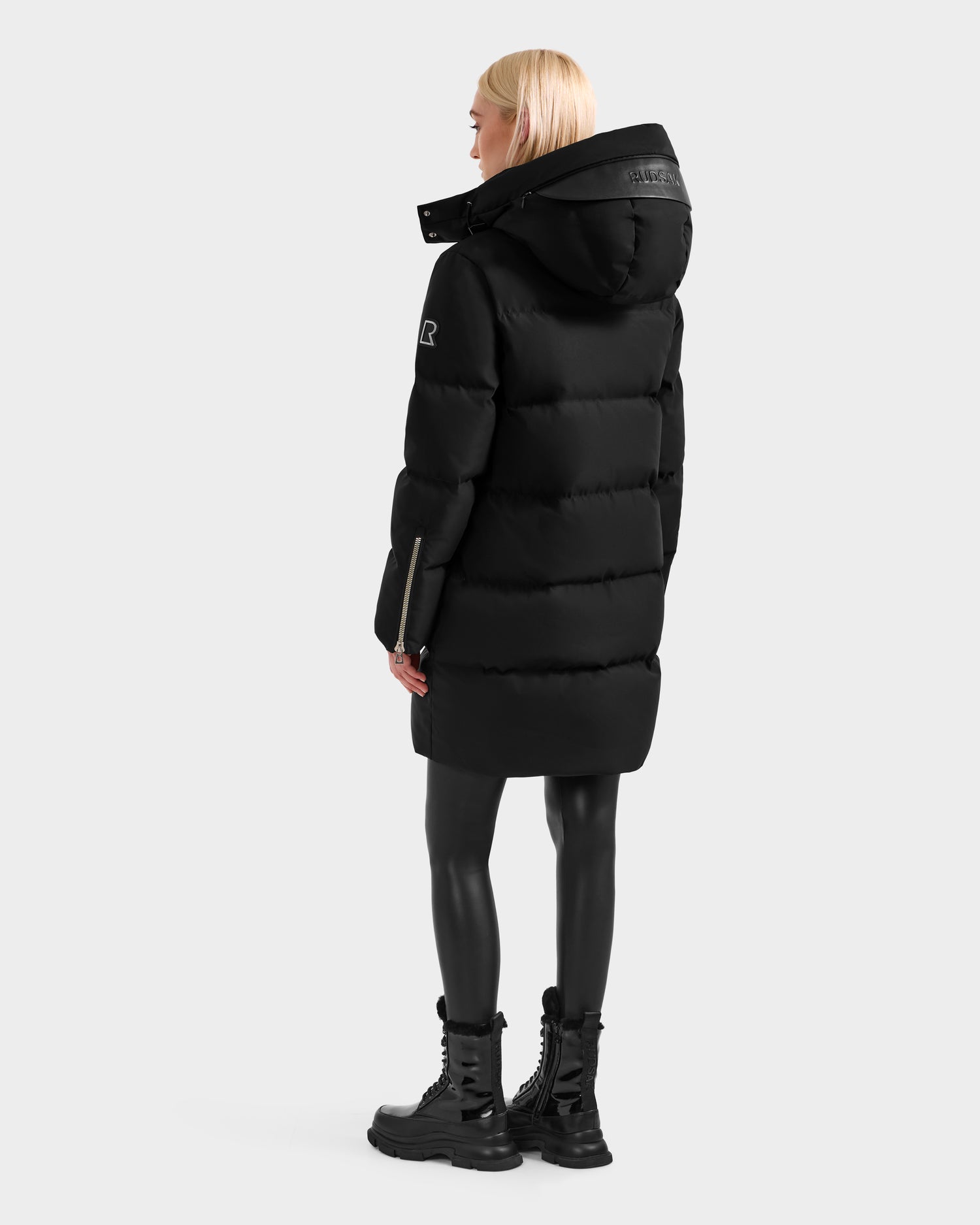 A person with blonde hair faces away, wearing the Rudsak JOELLE H - 8125602 Black water-resistant winter coat with a hood, black leggings, and black lace-up boots against a plain light gray background.