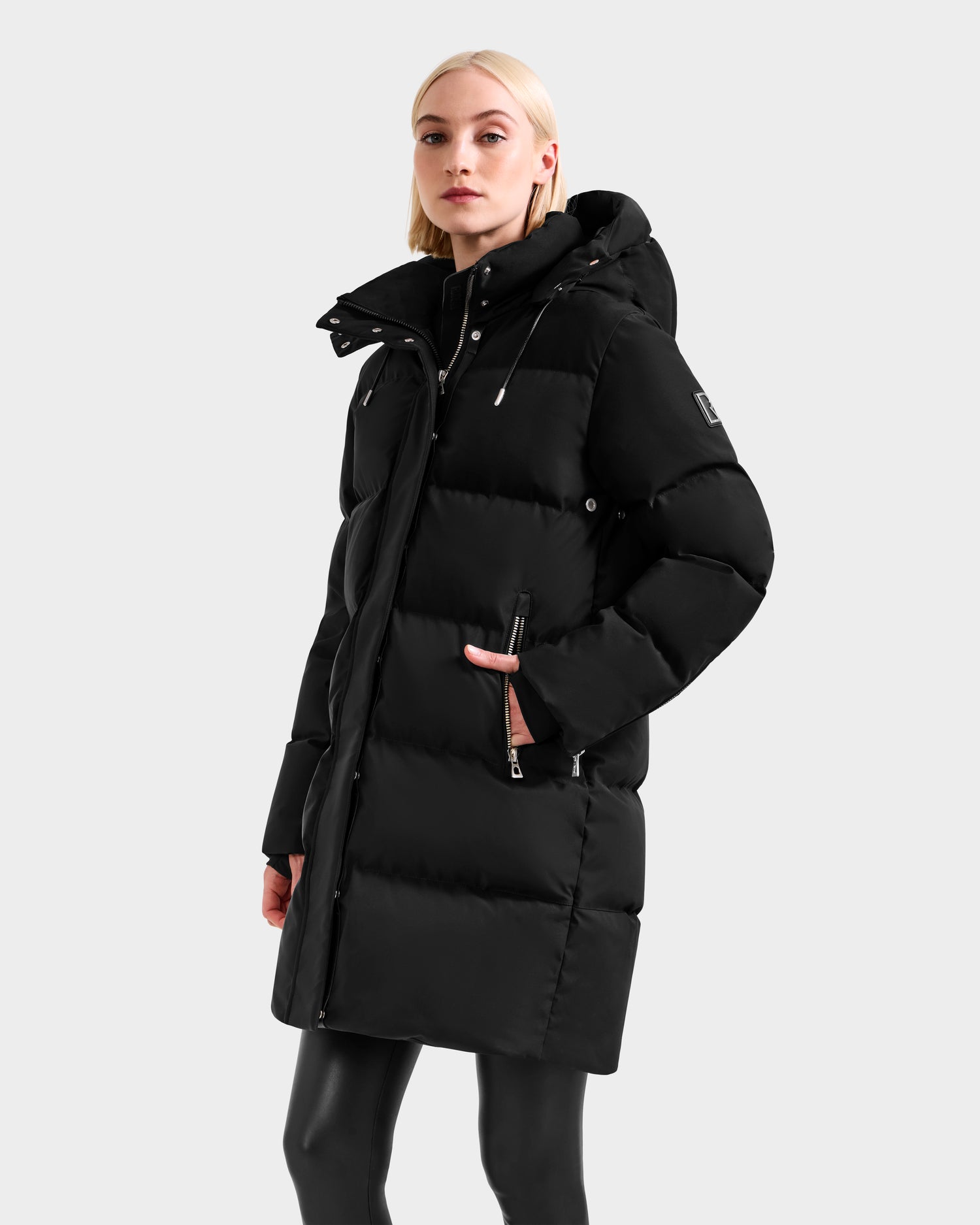 A woman with light blonde hair models the Rudsak JOELLE H - 8125602 Black, a long water-resistant winter coat with a hood and zippered pockets, against a plain light gray background.