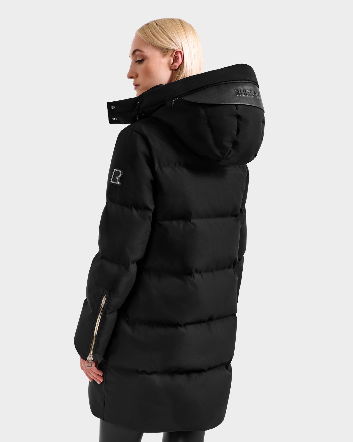 A woman with straight, blonde hair models the Rudsak JOELLE H - 8125602 Black long women's down puffer coat with hood and zipper details, standing against a light gray background and facing slightly away from the camera.