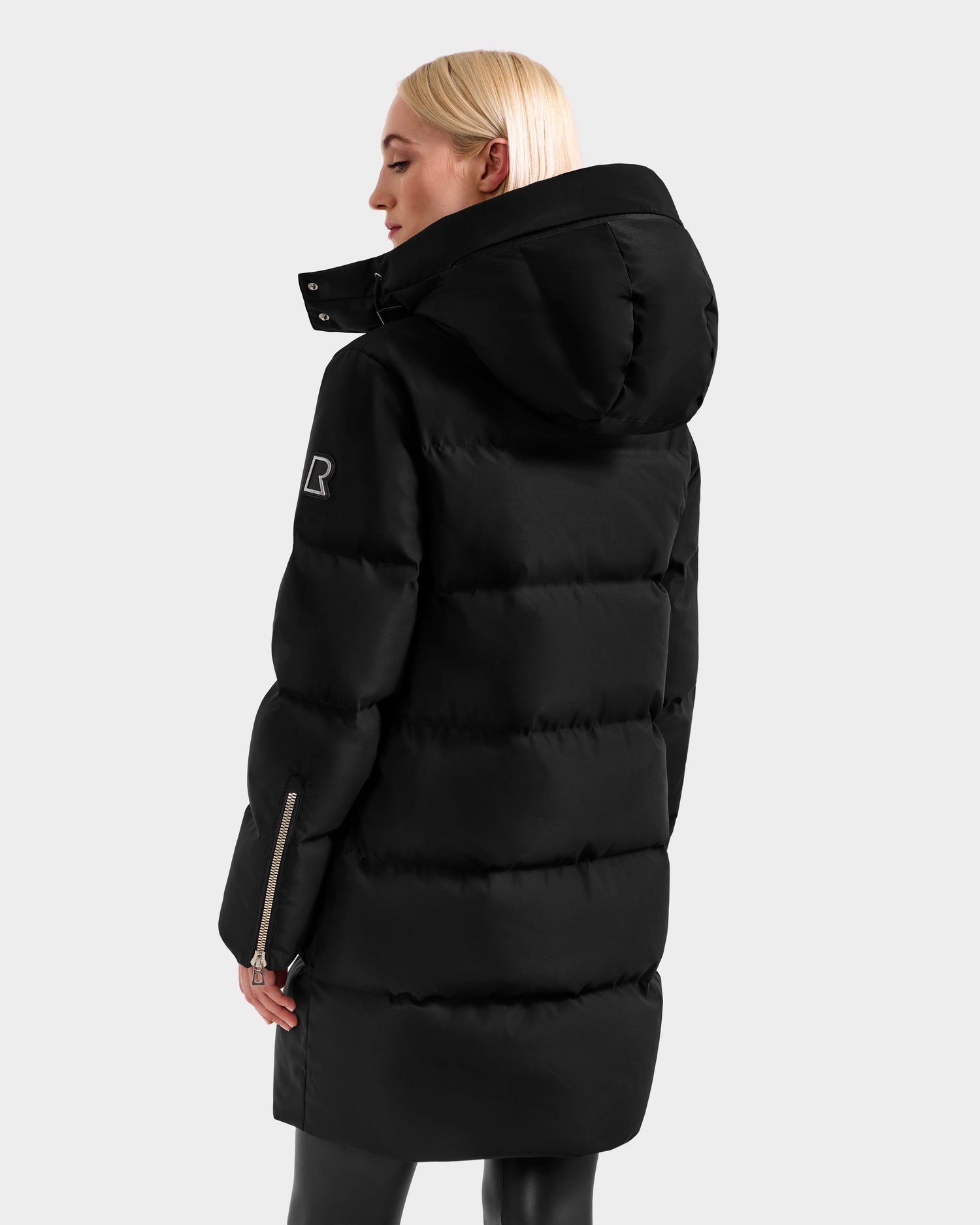 A person with straight blonde hair models the Rudsak JOELLE H - 8125602 Black women’s long down puffer winter coat with hood, water-resistant finish, zippered sleeve, and logo patch. Shown from the back against a light gray background.