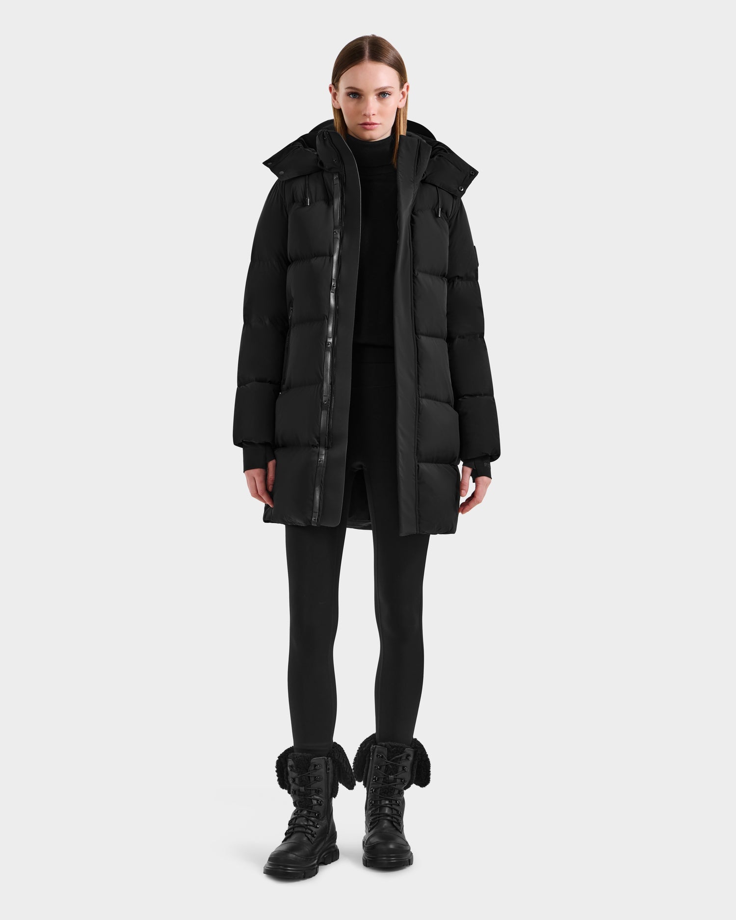 A person stands facing forward against a plain light background wearing the Rudsak JOELLE - 8124729 Black women's quilted down coat, styled with a black turtleneck, black leggings, and black lace-up boots.