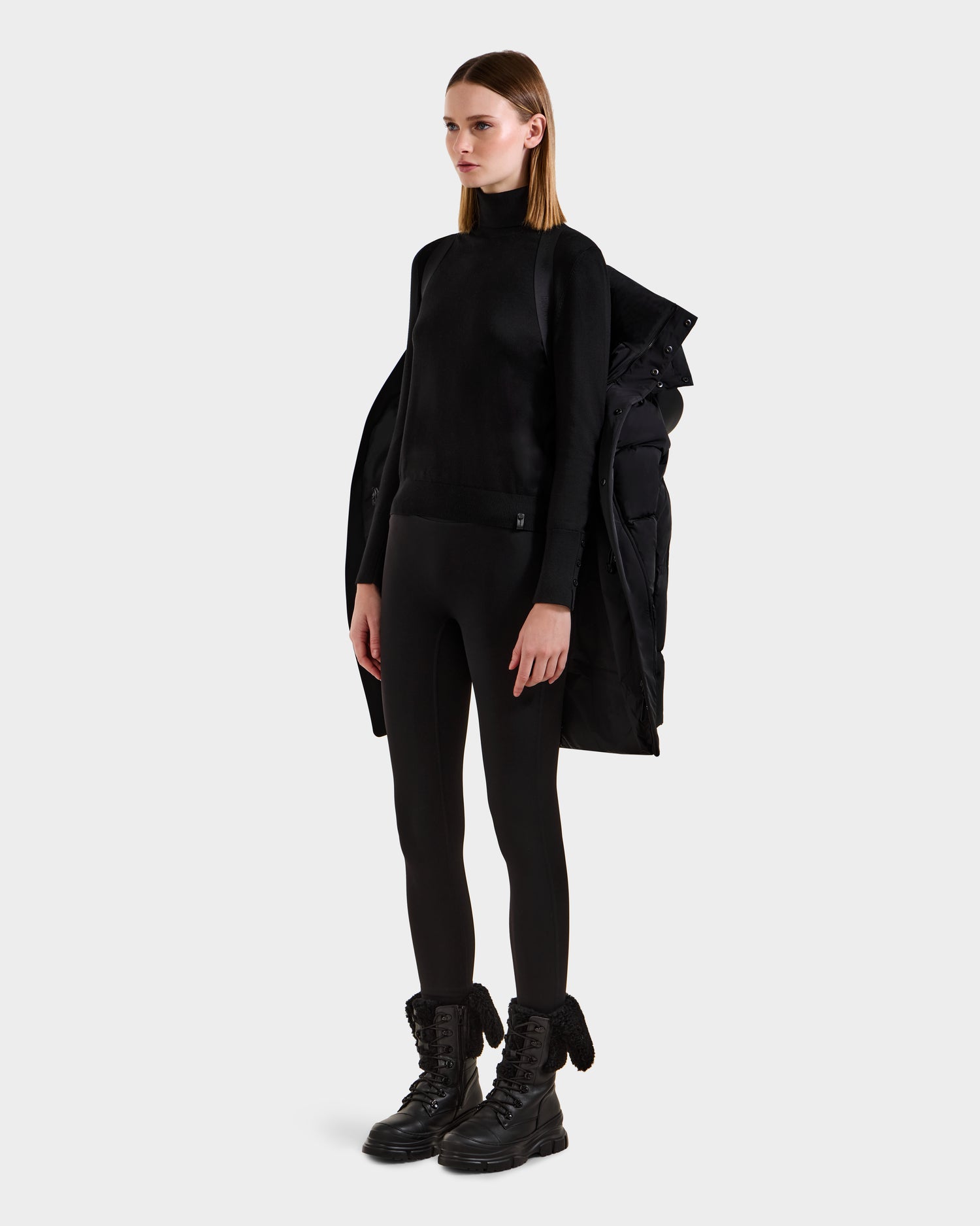 A woman stands against a plain background, wearing the Rudsak JOELLE - 8124729 Black quilted down coat draped over her shoulders, paired with a black turtleneck, leggings, and lace-up boots. She looks straight ahead with a neutral expression.
