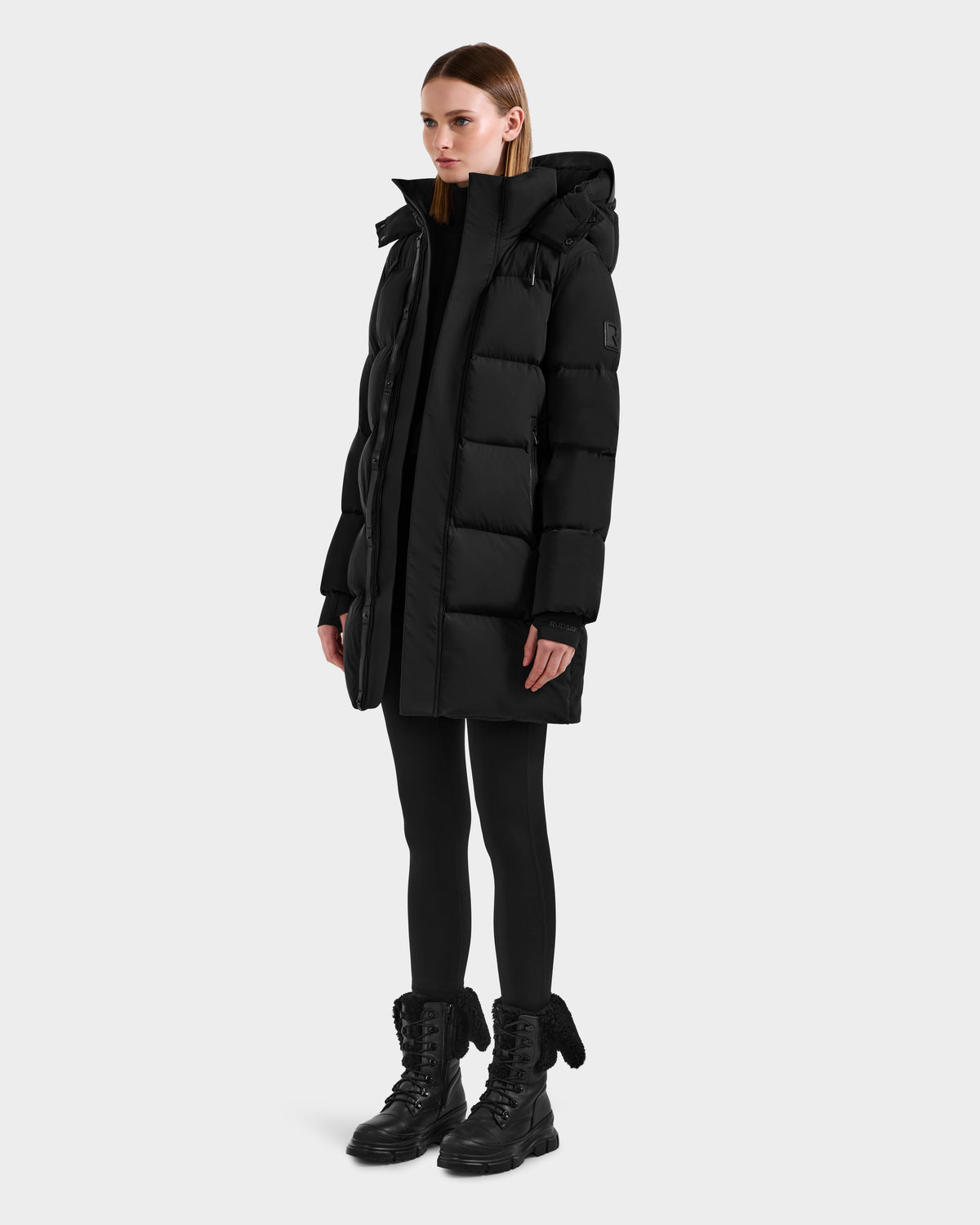 A woman stands in the Rudsak JOELLE - 8124729 Black quilted down coat with a hood, paired with black leggings and lace-up boots. She faces slightly to the side against a plain white background, ready for winter in this stylish coat.