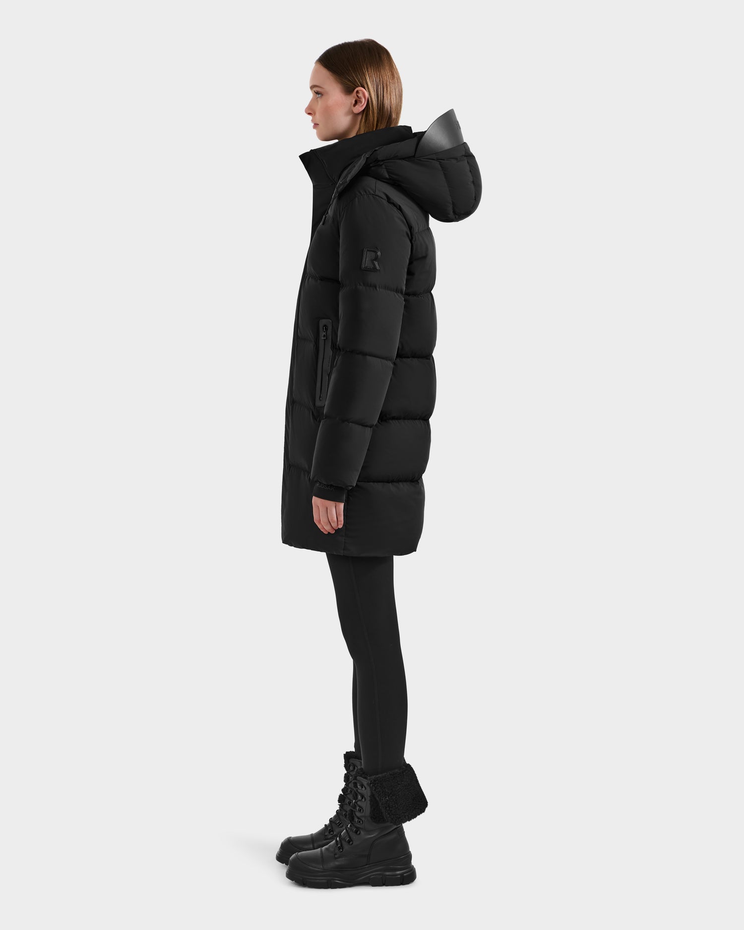 A woman in profile wears the Rudsak JOELLE - 8124729 Black quilted down coat with high collar and hood, paired with black leggings and lace-up boots, set against a plain light gray background.