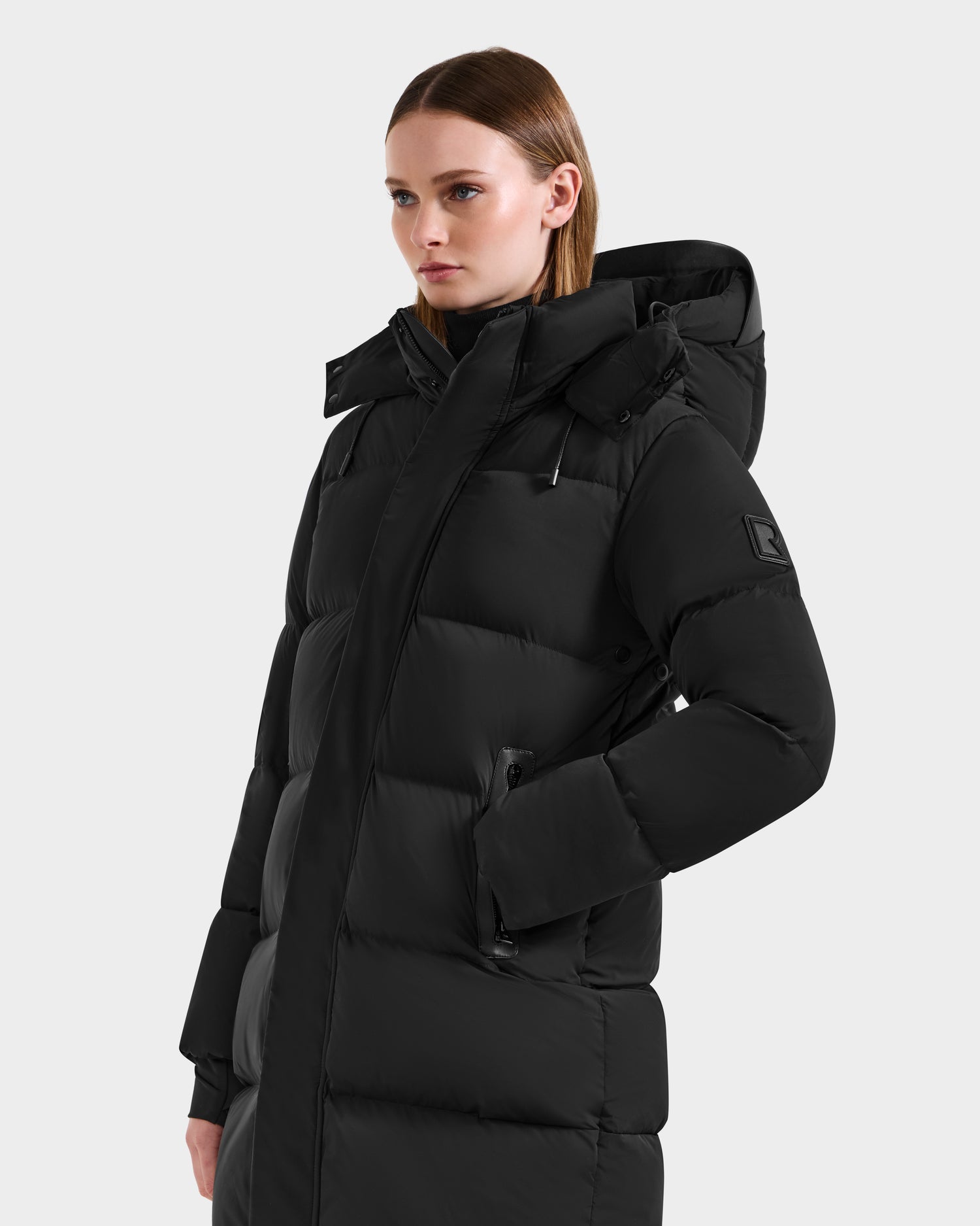 A woman with straight light brown hair models the Rudsak JOELLE - 8124729 Black quilted down coat with hood and zippered pockets, standing against a plain light gray background.