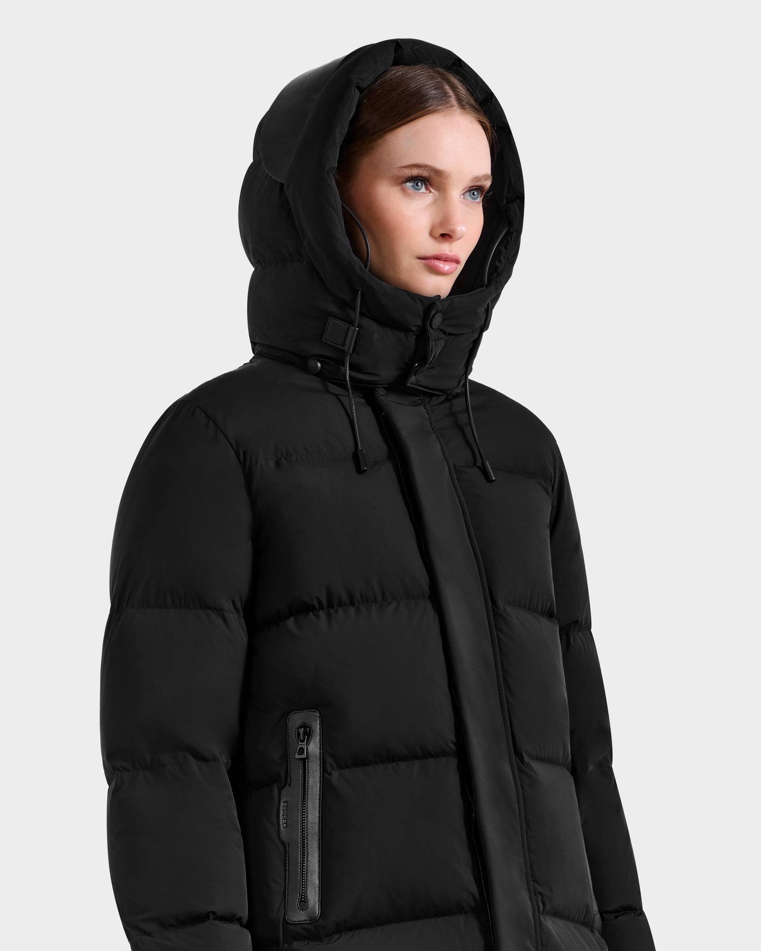 A woman models the Rudsak JOELLE - 8124729 Black puffy quilted down coat with zippered pockets, standing against a light grey background—an ideal winter staple.