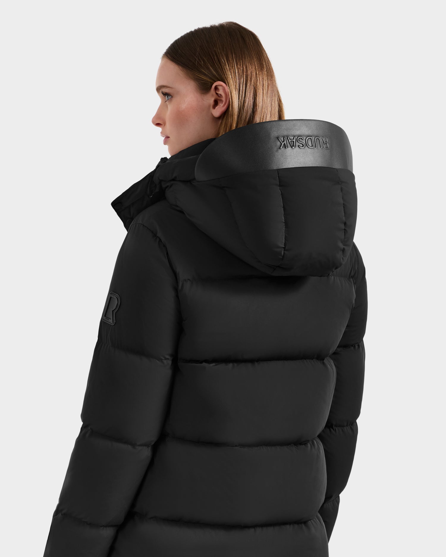 A woman with straight brown hair wears the Rudsak JOELLE - 8124729 Black quilted down coat with hood, featuring RDS-certified down and logo details on the collar and sleeve, standing sideways against a plain light background.