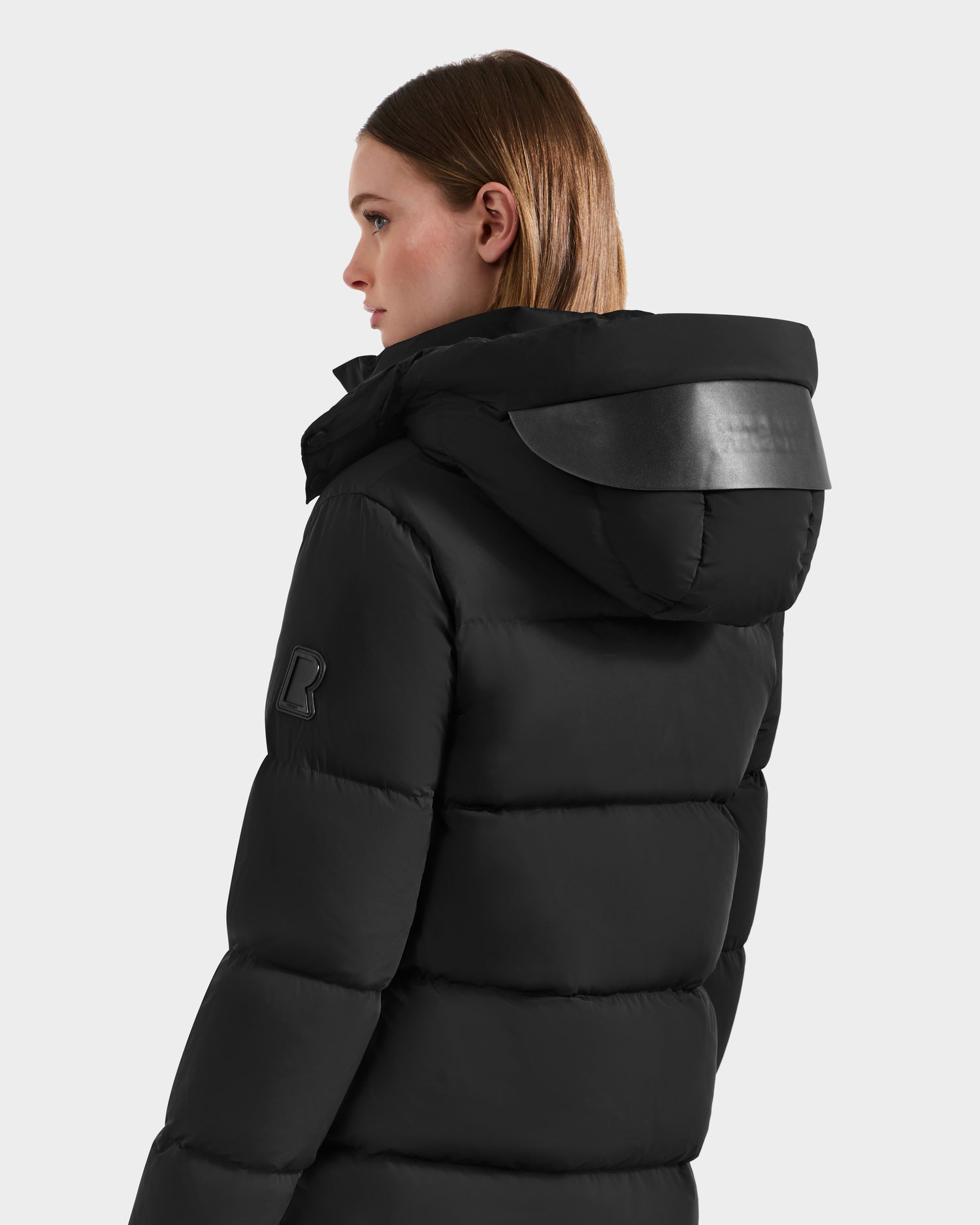 A woman with straight, light brown hair wears the Rudsak JOELLE - 8124729 Black women's quilted down coat with a hood, facing away against a light background to showcase this RDS-certified winter staple.