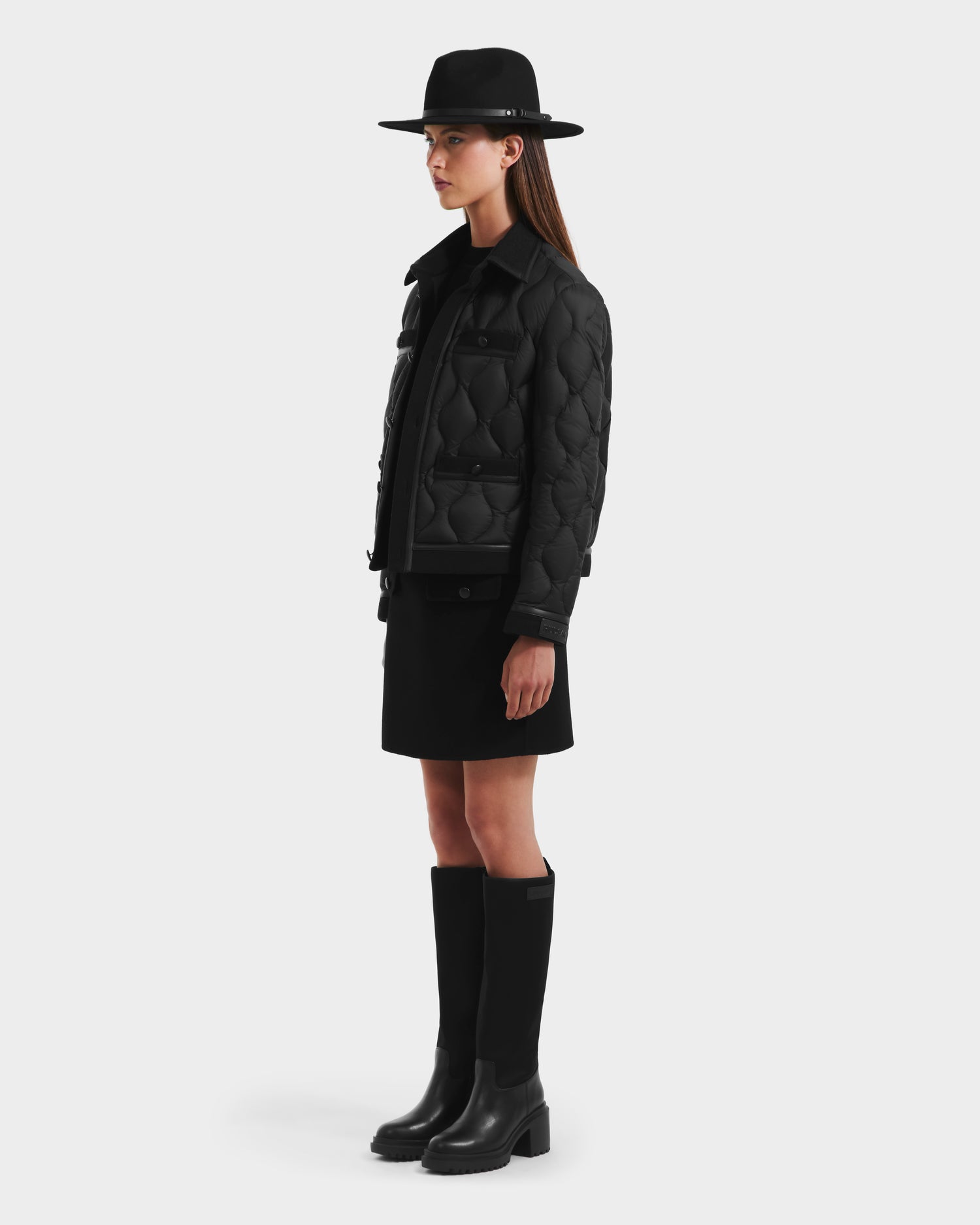 A woman in profile against a white background models the Rudsak JOLIE - 8125036 Black quilted down jacket, paired with a black wide-brimmed hat, black skirt, and knee-high boots for chic style and RDS-certified down warmth.