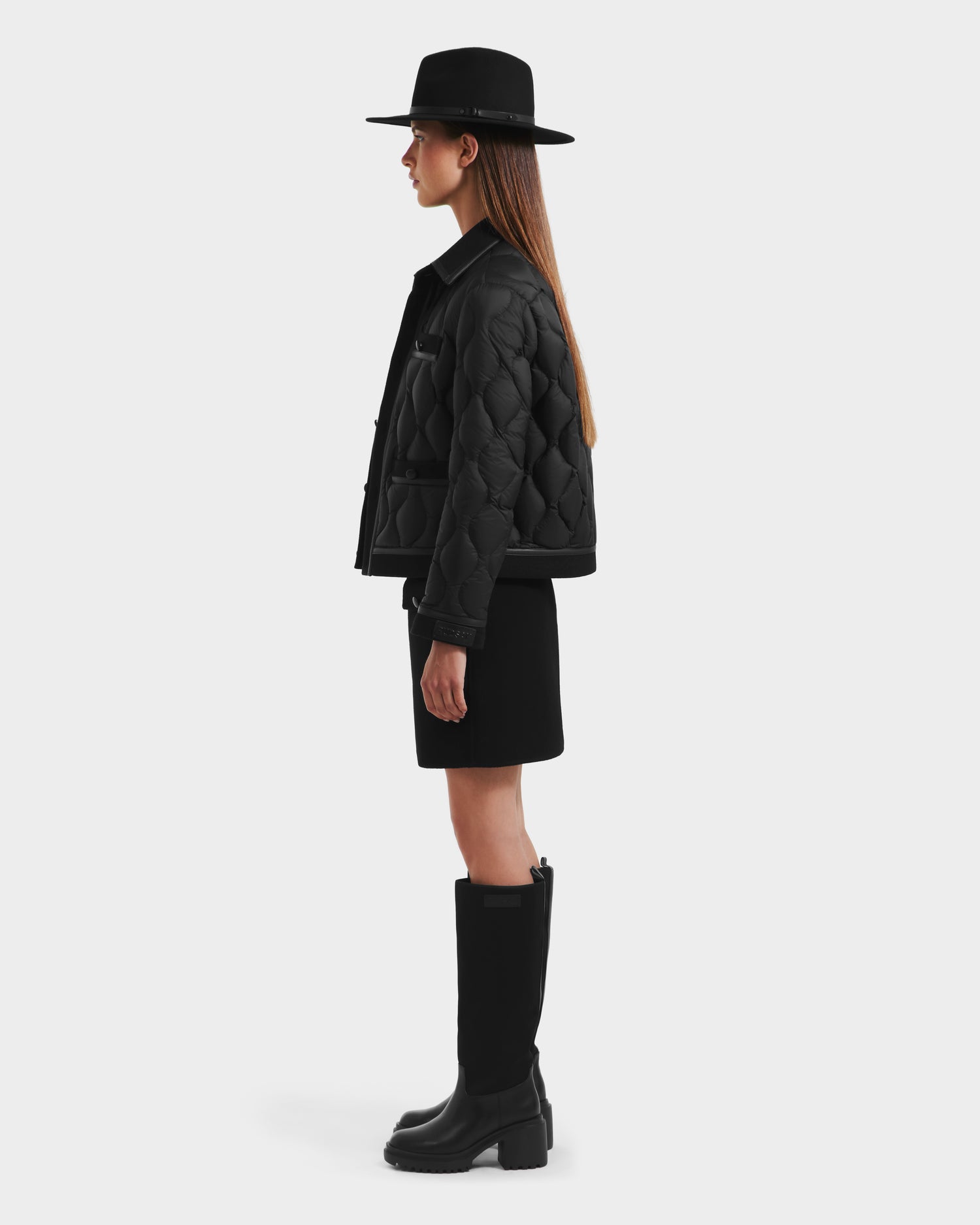 A woman stands in profile wearing the Rudsak JOLIE - 8125036 Black quilted down jacket with premium RDS-certified down, paired with a black knee-length skirt, tall black boots, and a wide-brimmed hat against a plain light background.