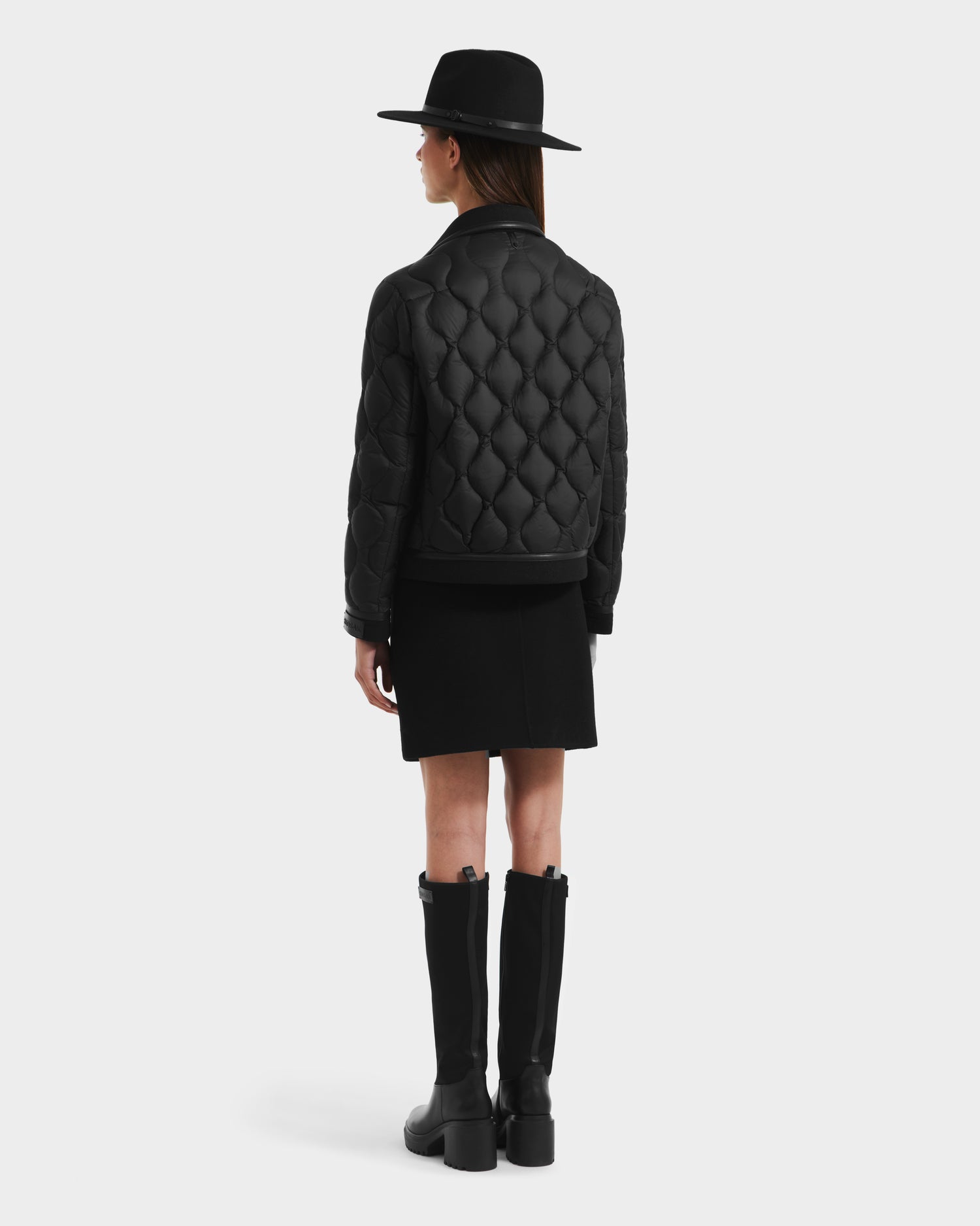 A woman faces away, wearing the Rudsak JOLIE - 8125036 Black women’s quilted down jacket with RDS-certified fill, styled with a black skirt, tall boots, and a wide-brimmed hat against a plain light background.