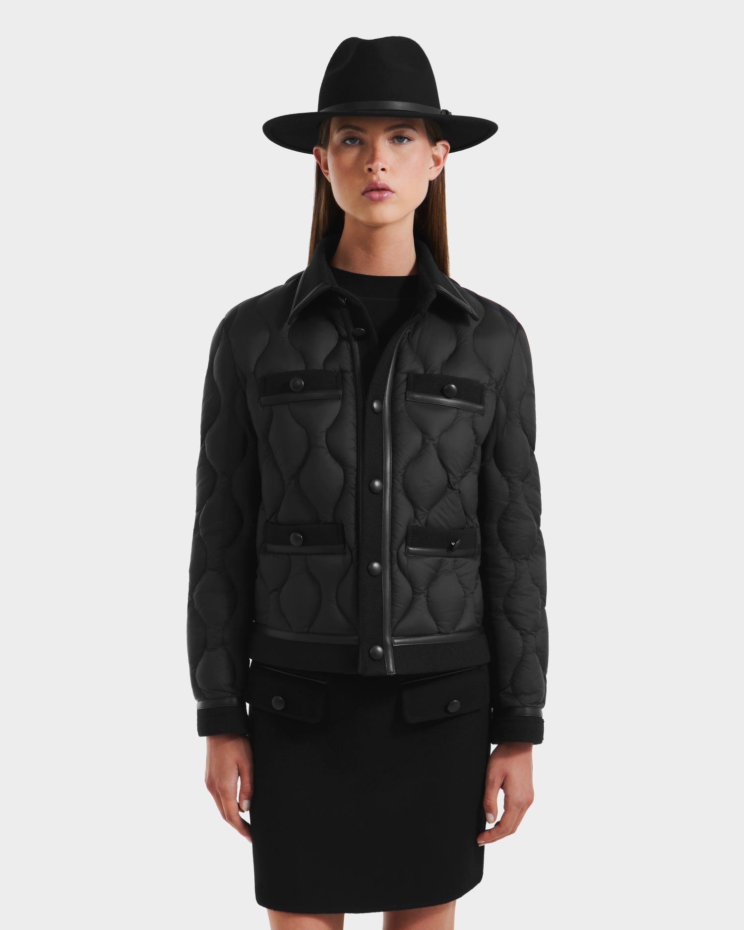 A woman in the Rudsak JOLIE - 8125036 Black quilted down jacket with snap buttons and pockets, a wide-brimmed hat, and a black skirt stands against a plain light gray background.