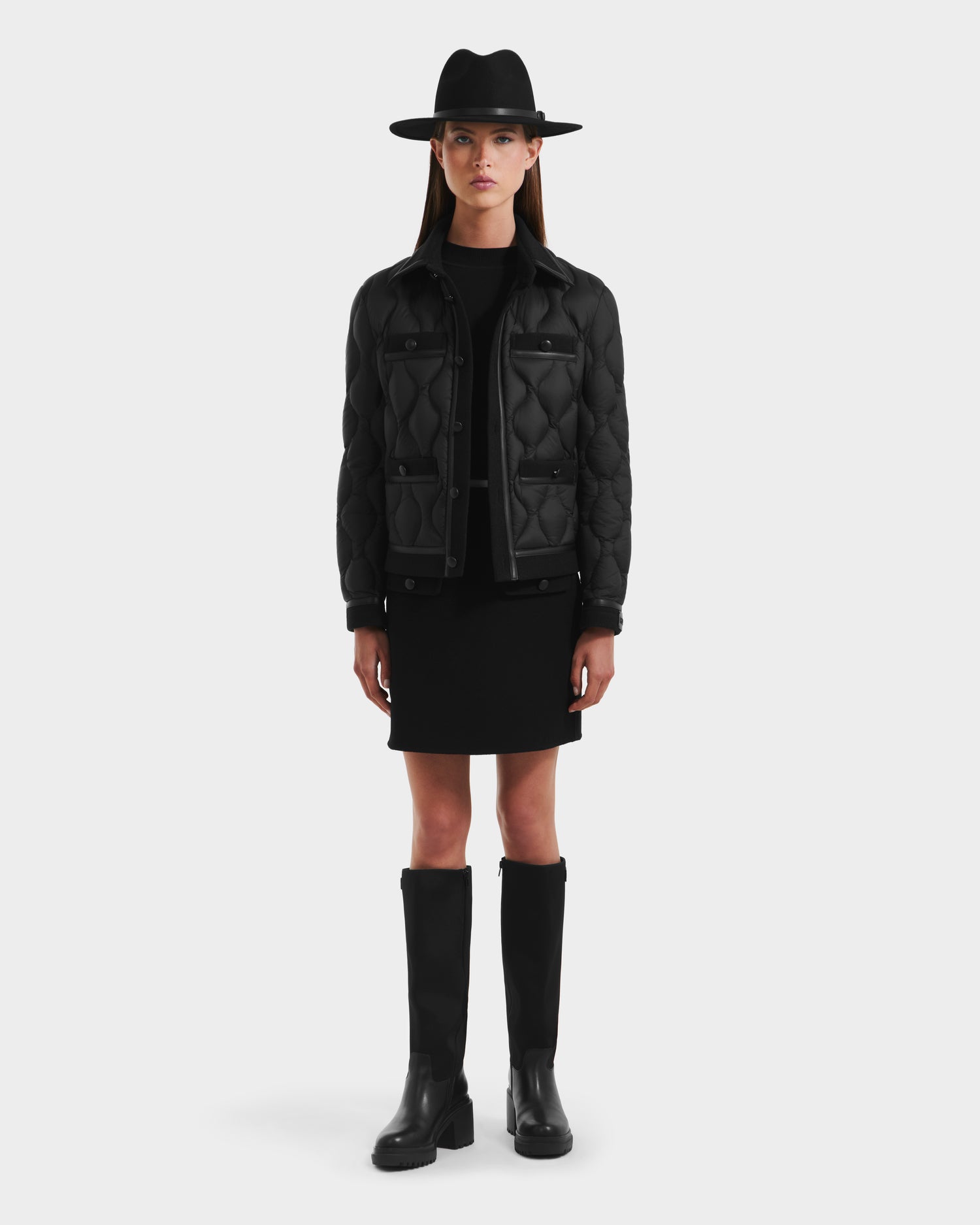 A woman stands facing forward in a Rudsak JOLIE - 8125036 Black quilted down jacket, paired with a black skirt, tall black boots, and a wide-brimmed hat against a plain light background.