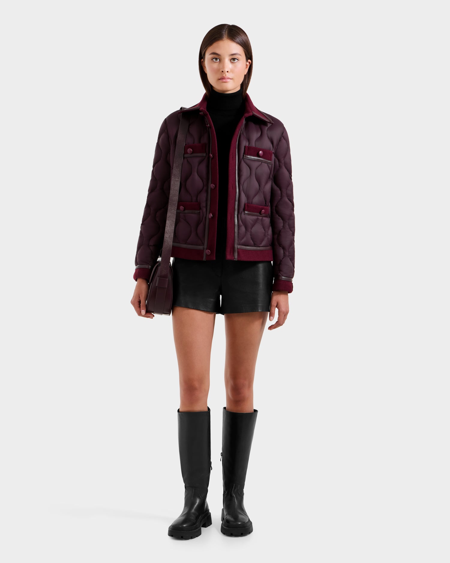 A woman faces forward against a light background, wearing the Rudsak JOLIE - 8125036 Bordeaux women’s quilted light down jacket with vegan leather-covered buttons, paired with a black turtleneck, shorts, knee-high boots, and a matching maroon shoulder bag.