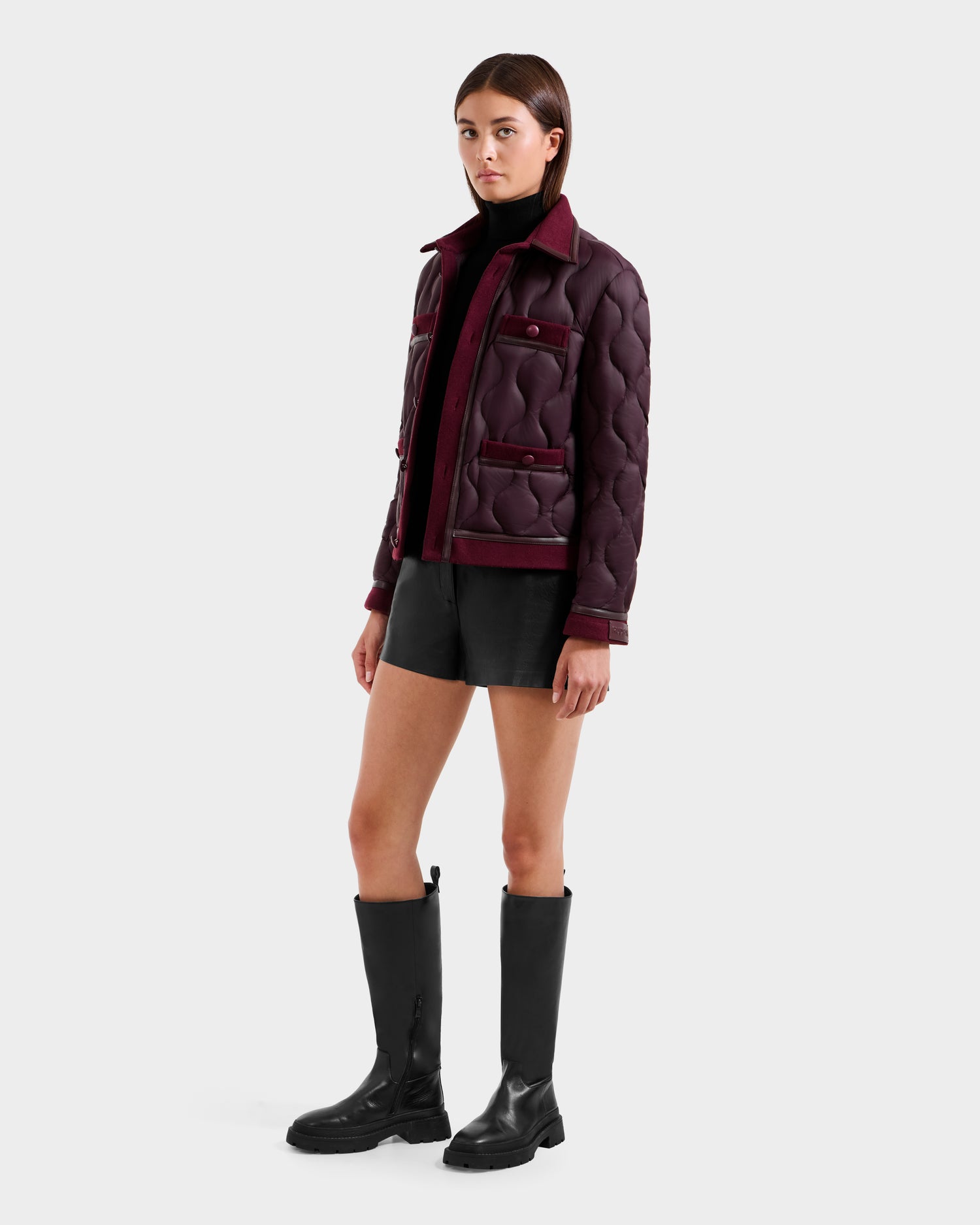 A person with straight brown hair and a neutral expression wears the Rudsak JOLIE - 8125036 Bordeaux women’s quilted light down jacket, styled with a black turtleneck, black shorts, and knee-high black boots against a plain background.