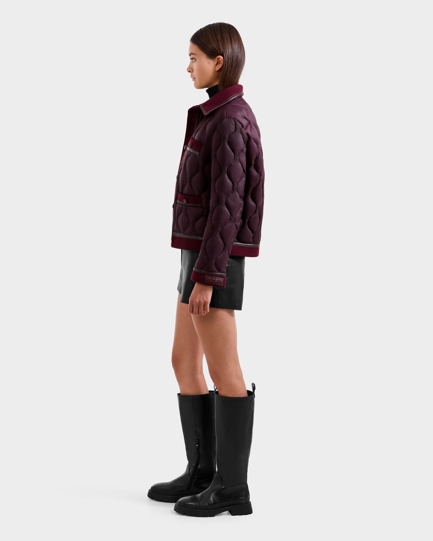 A woman in profile wears the Rudsak JOLIE - 8125036 Bordeaux quilted jacket, a black leather mini skirt, and knee-high black boots against a plain light background.