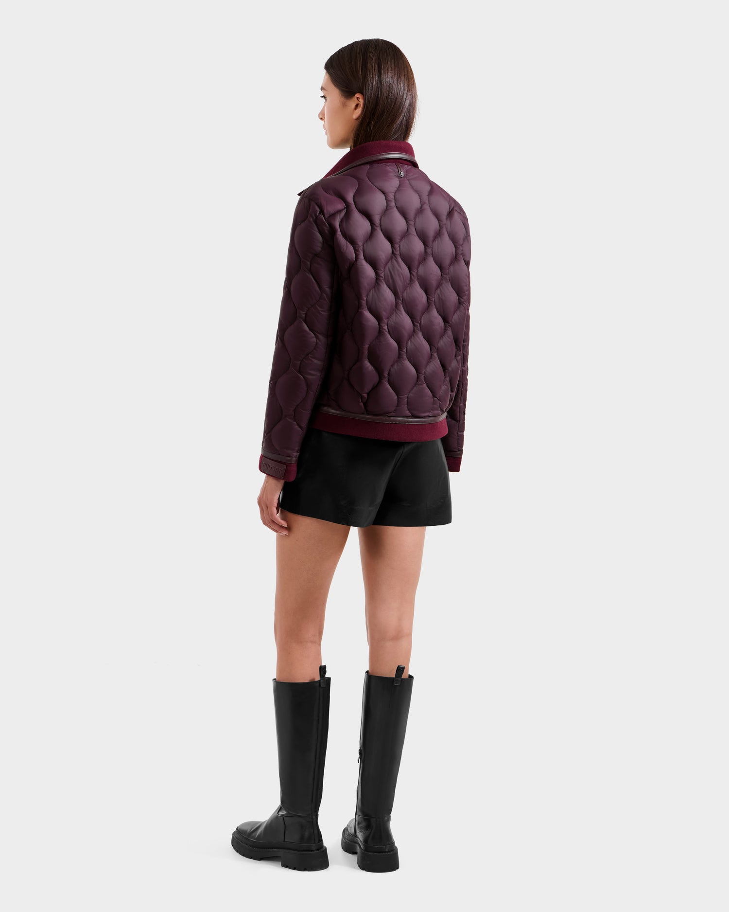 A woman faces away, wearing Rudsak's JOLIE - 8125036 Bordeaux quilted light down jacket with vegan leather-covered buttons, black shorts, and knee-high black boots against a plain light background.
