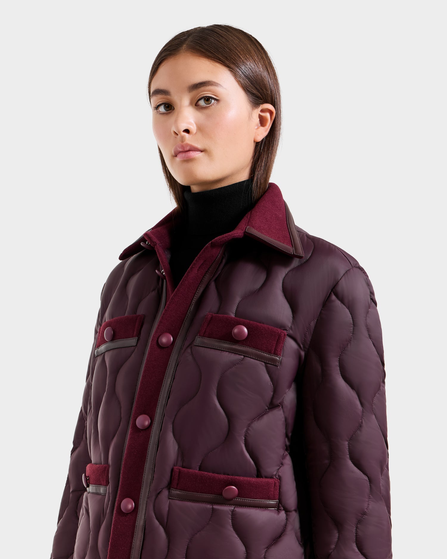A woman with straight brown hair wears the Rudsak JOLIE - 8125036 Bordeaux quilted down jacket featuring buttoned pockets, RDS-certified down, and a high collar over a black turtleneck against a plain light gray background.