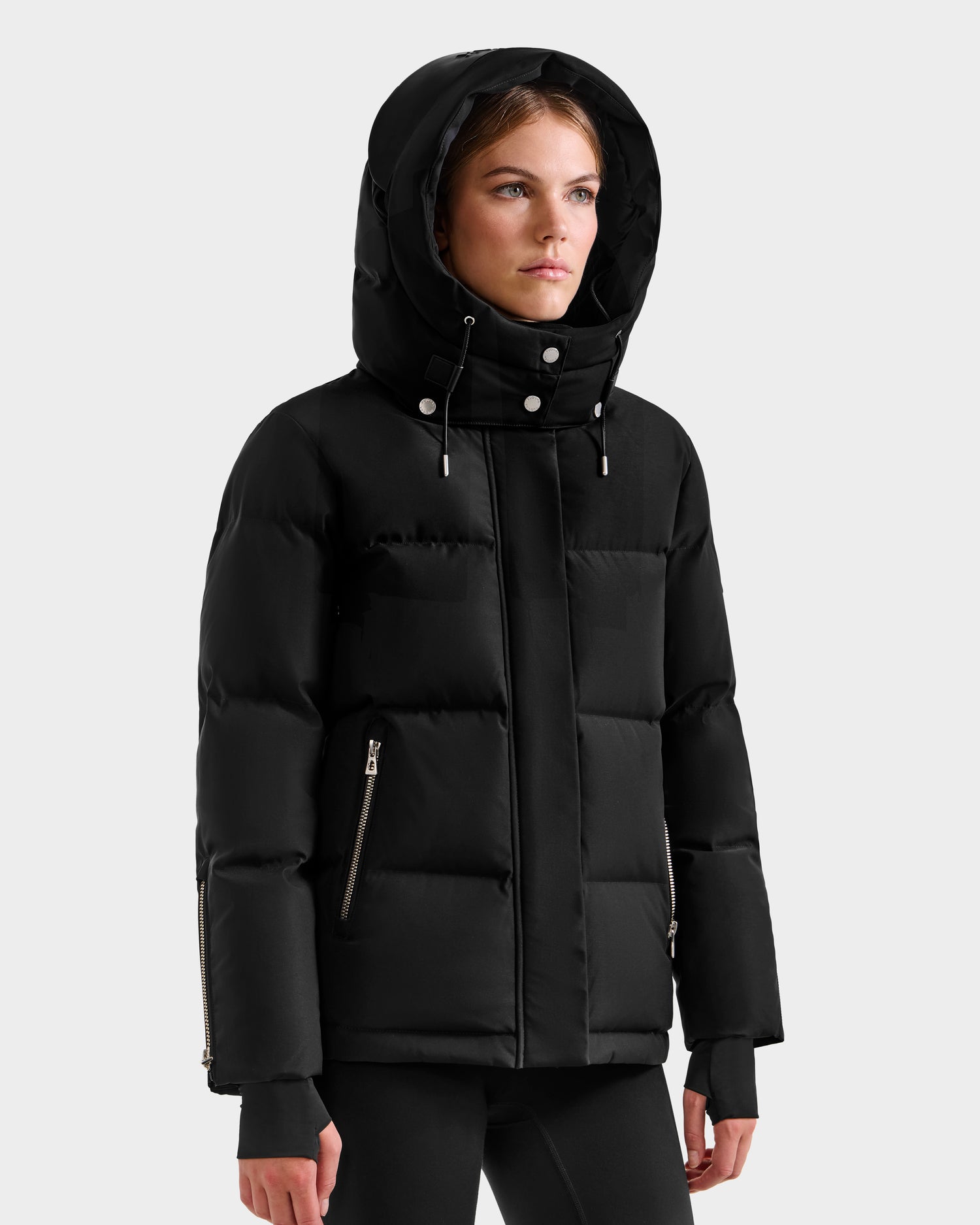A woman stands against a plain light gray background wearing the Rudsak JOON H - 8125586 Black insulated quilted jacket, featuring a hood, zippered pockets, and side vents.